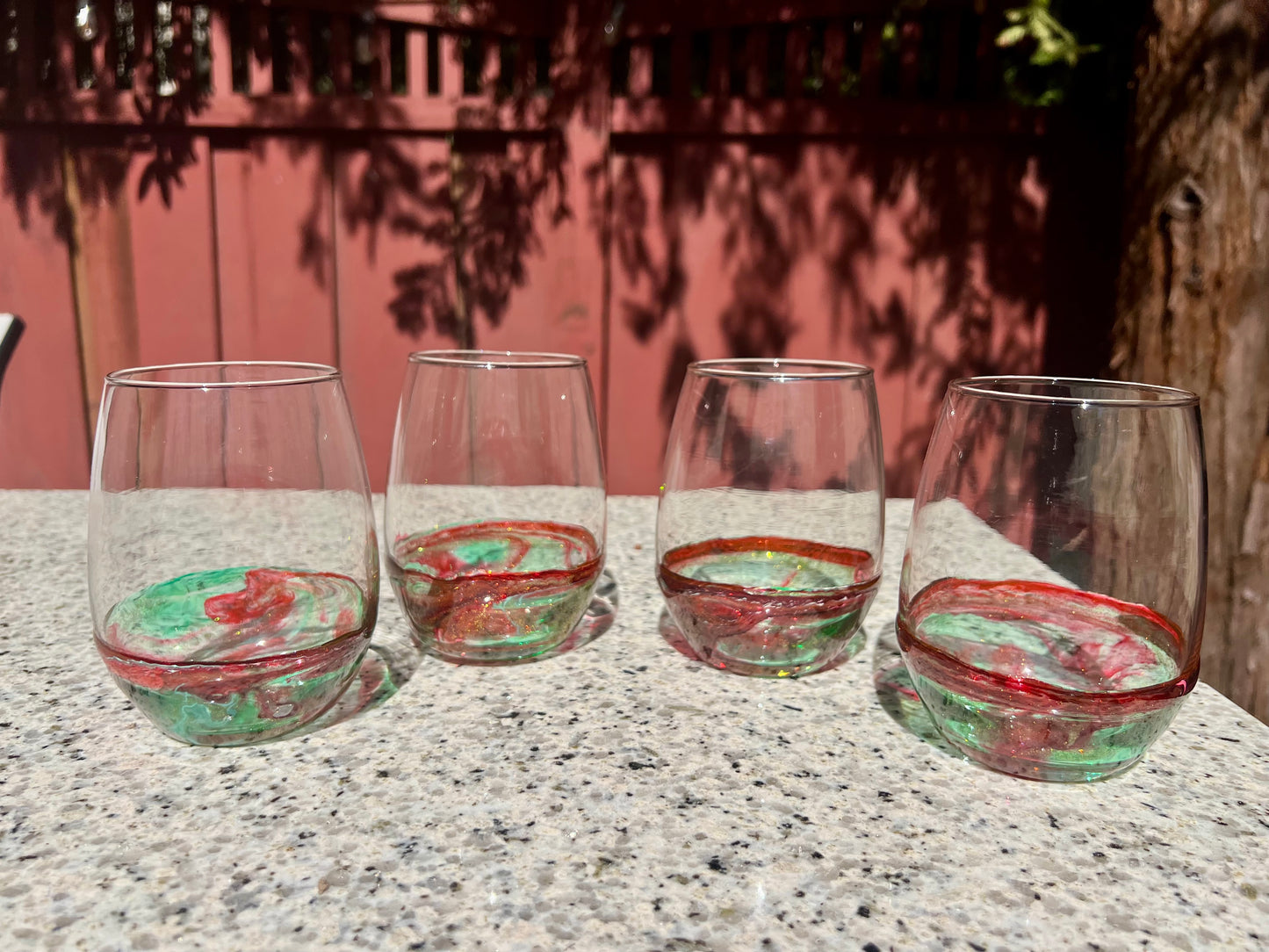 Four wine glasses with red and green swirls are arranged on a granite countertop, with a red fence and trees in the background.