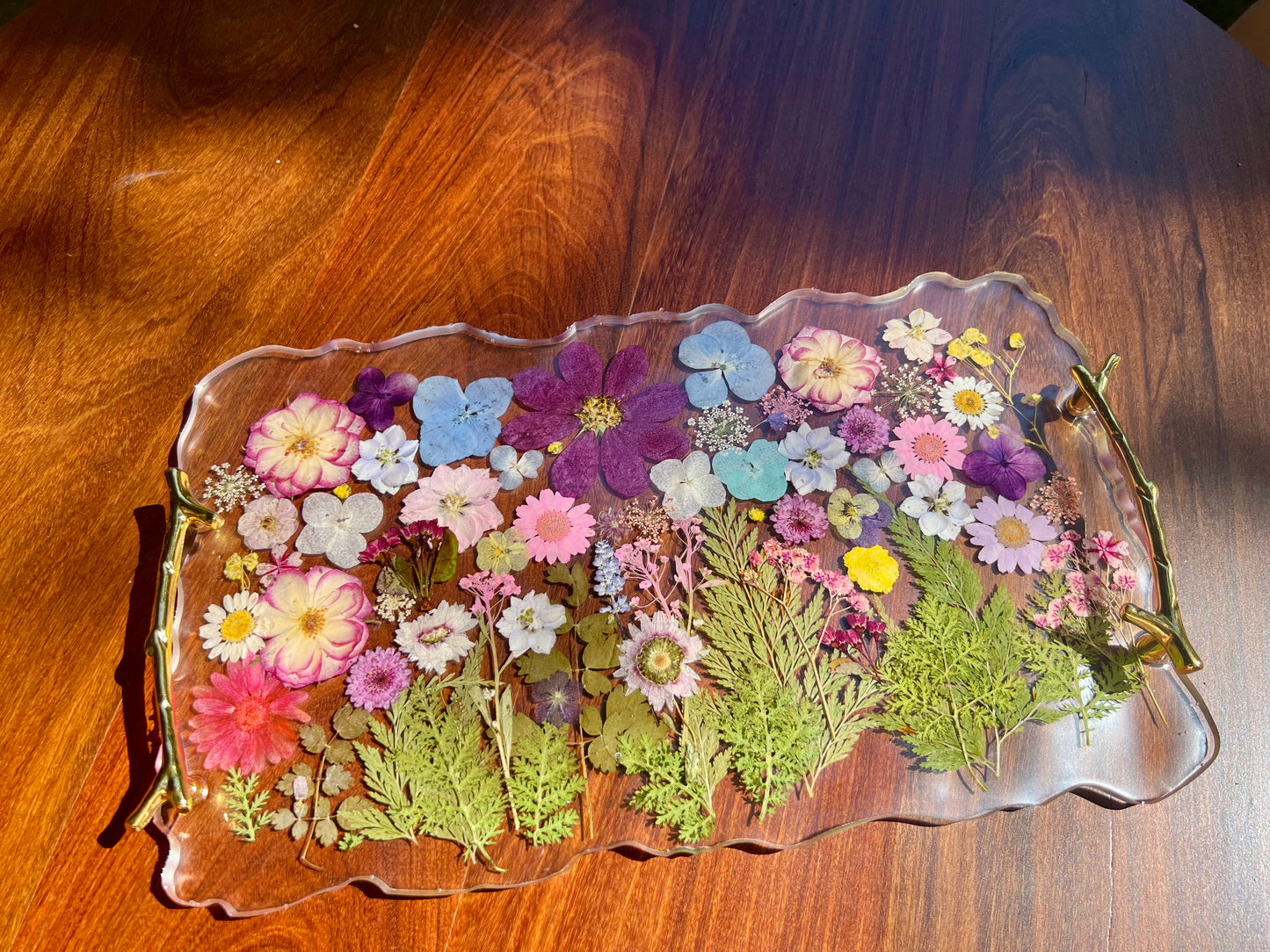 A clear glass tray filled with a variety of colorful flowers and leaves, including pink, purple, yellow, and green flowers, as well as green leaves, arranged on a wooden surface.