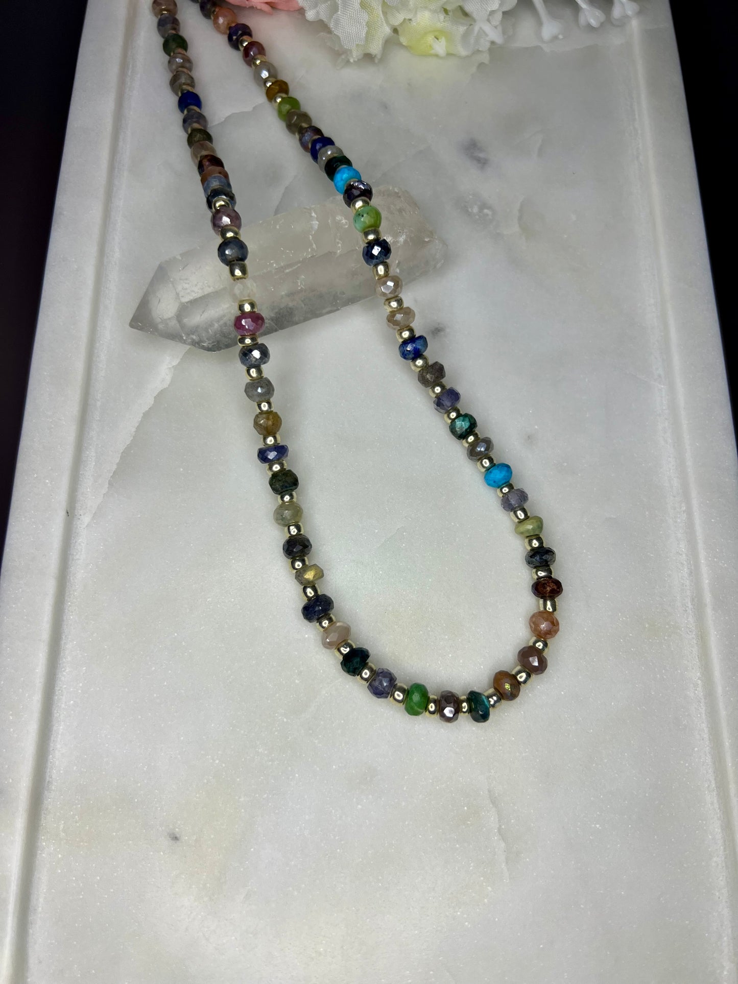 A colorful beaded necklace with various colored beads, including blue, green, purple, and gold, is displayed on a white marble surface.