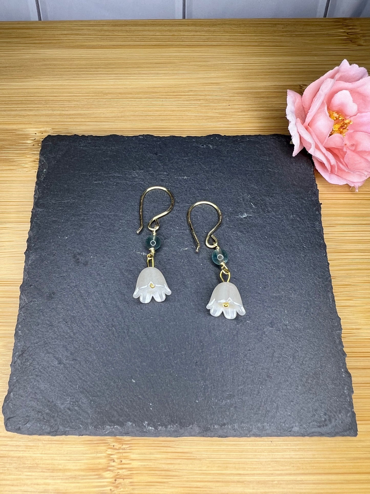 A pair of white flower-shaped earrings with green accents, hanging from gold hooks, are displayed on a black slate surface. A pink flower is also present in the background.