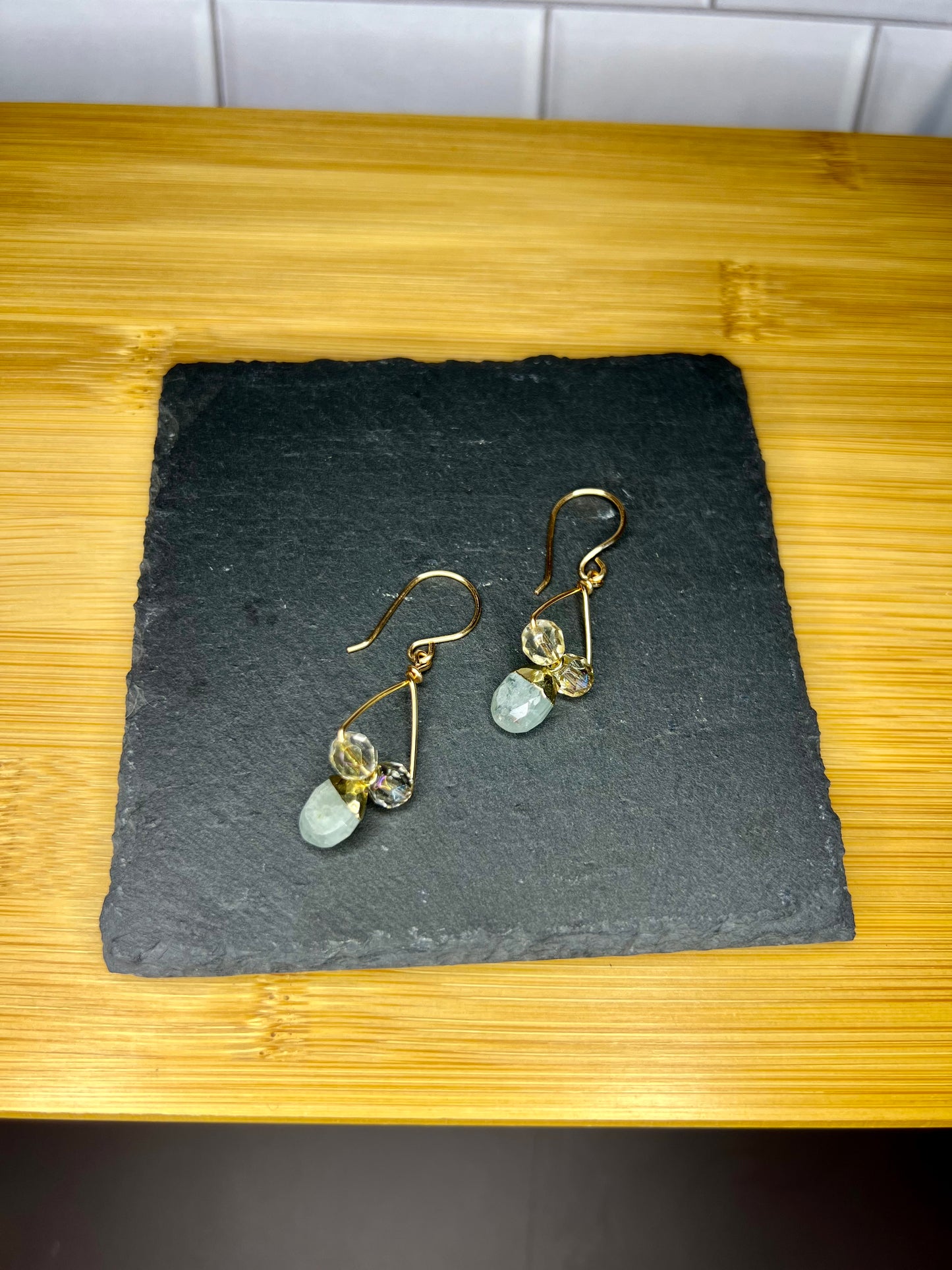 A pair of gold earrings with blue and green stones hanging from them, displayed on a black slate or slate board.
