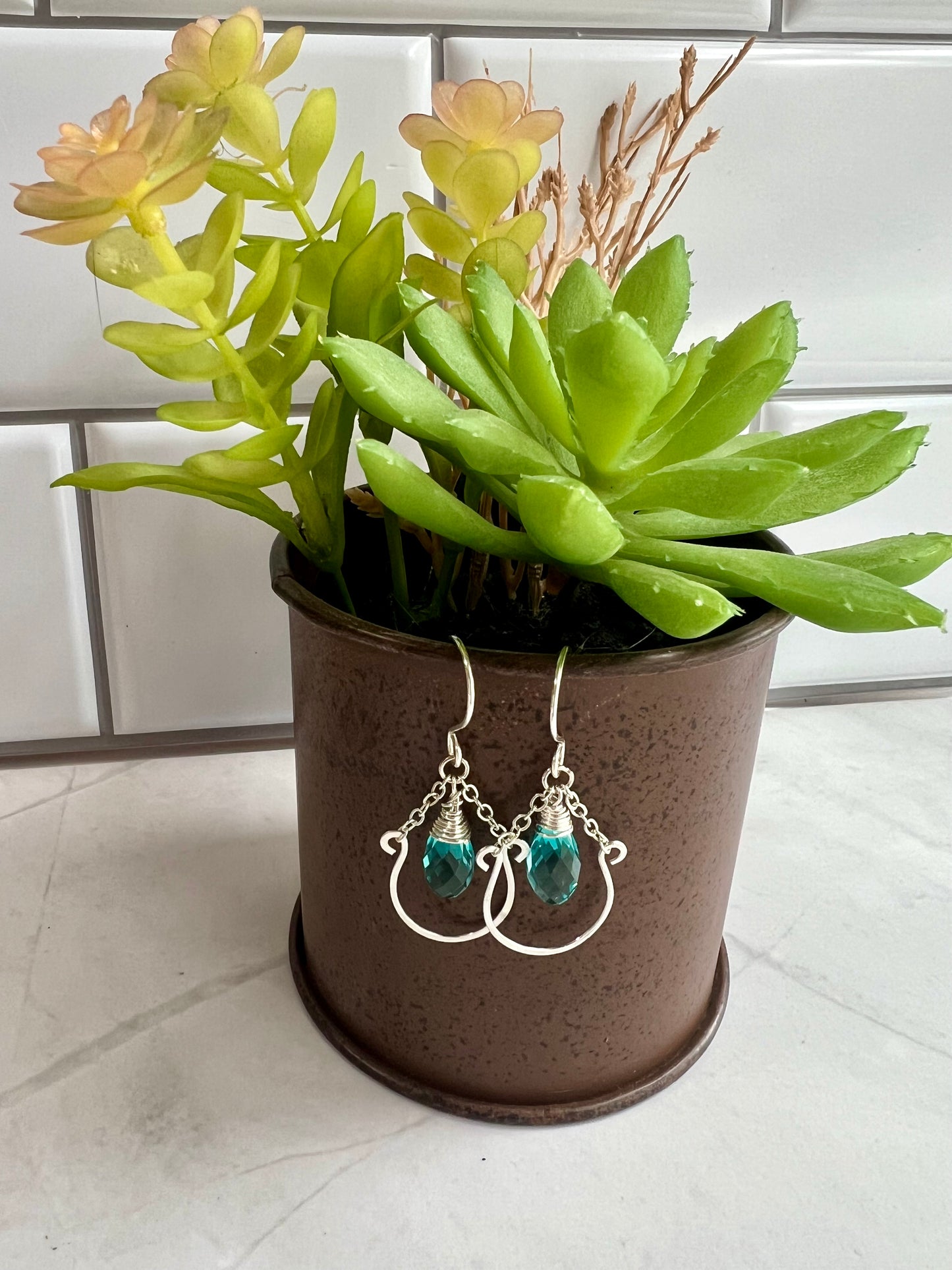 A pair of earrings with turquoise stones is displayed in a brown pot, surrounded by a variety of green plants.