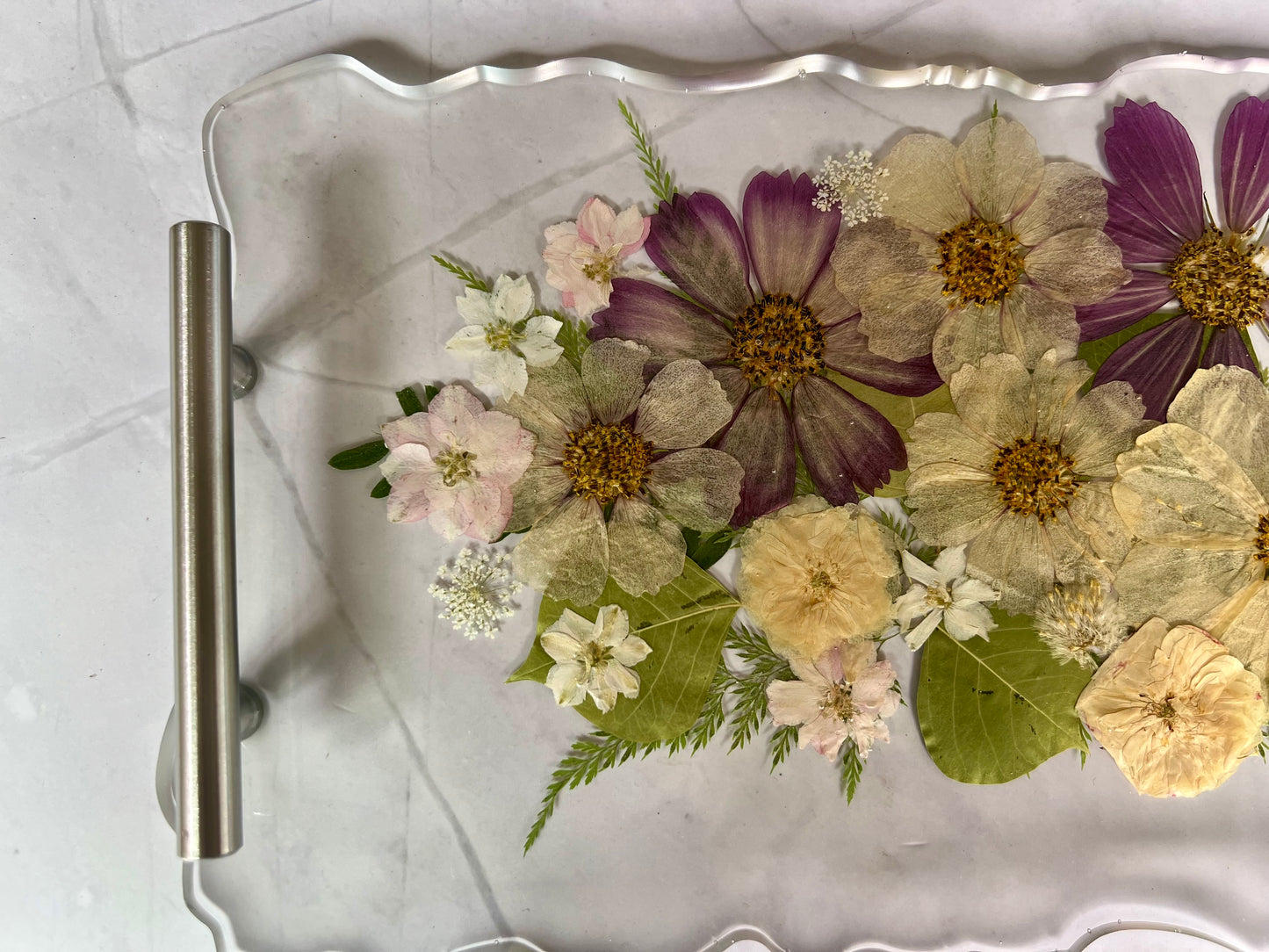 A clear glass tray with a metal handle, displaying a variety of colorful dried flowers and leaves arranged on it.