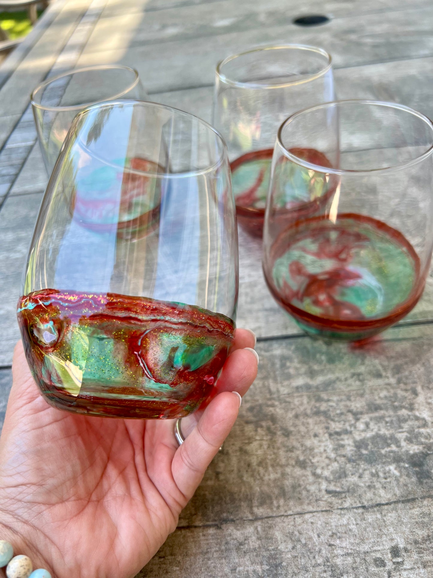 A hand holding a glass with a red and green liquid, with other glasses visible in the background on a wooden surface.