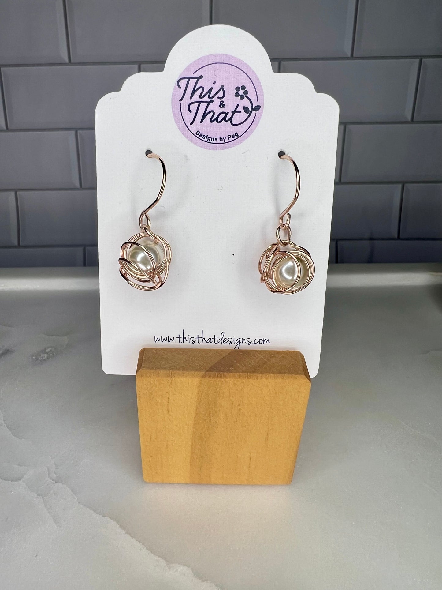 This image shows a pair of earrings displayed on a white card stand. The earrings are made of metal and have a unique, twisted design. The card stand is made of wood and has a website address printed on it.