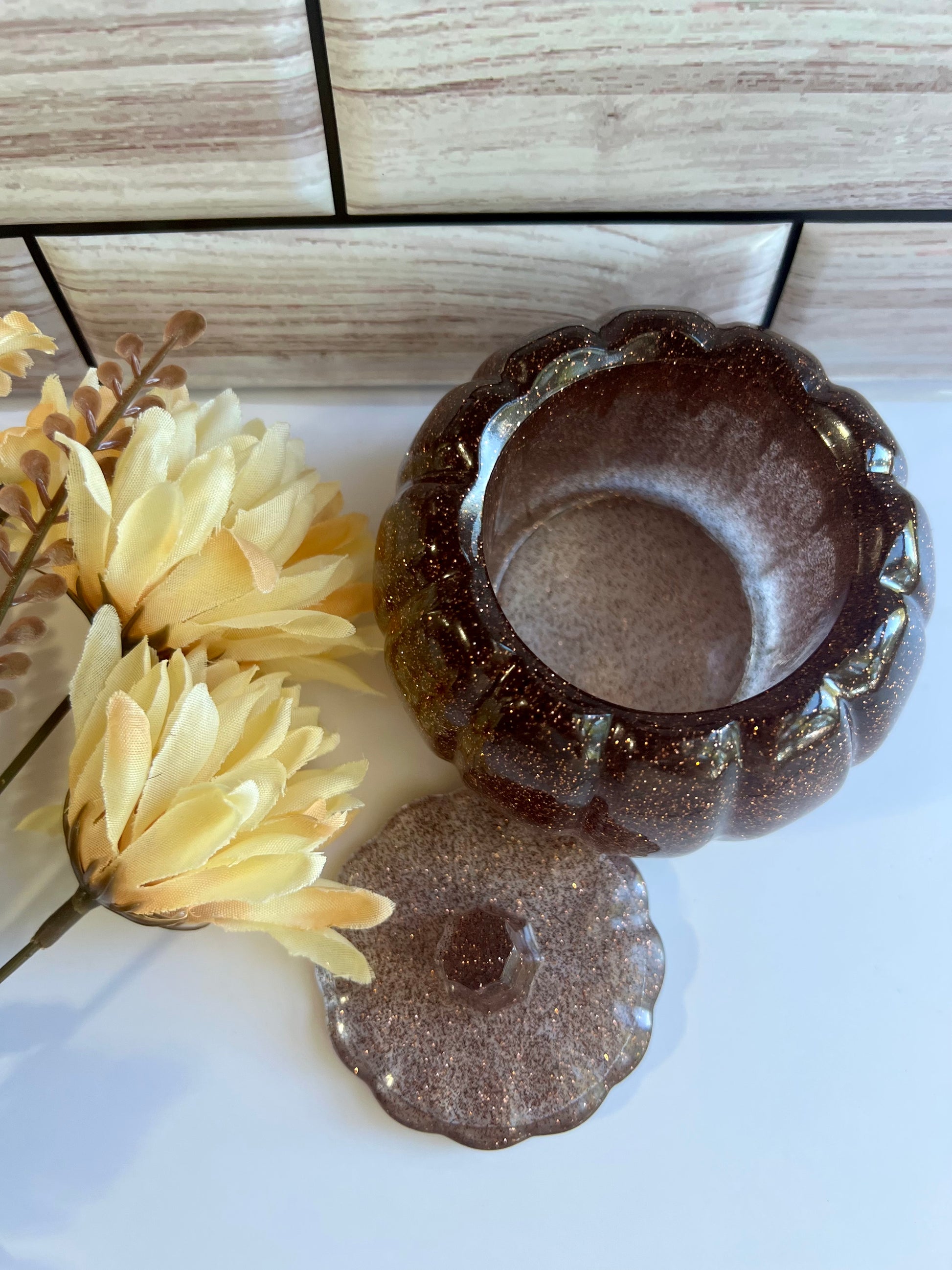 a brown glass bowl with a gold-colored lid, placed on a white surface next to a bunch of yellow flowers.