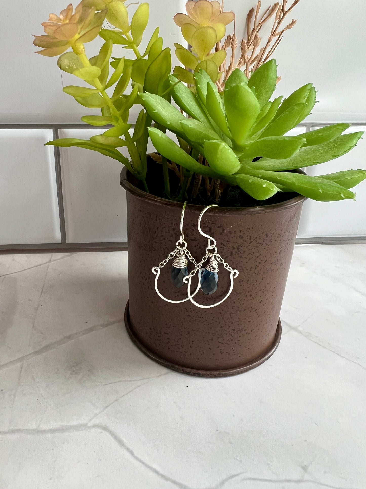 A pair of silver earrings with blue stones is displayed in a brown pot, placed on a white countertop.