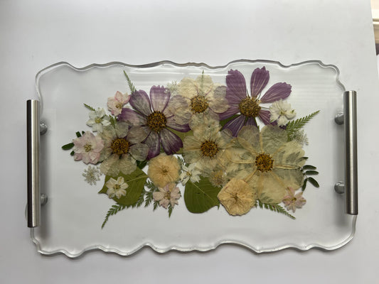 a clear glass tray with a floral arrangement of various flowers, including purple and white flowers, along with green leaves.