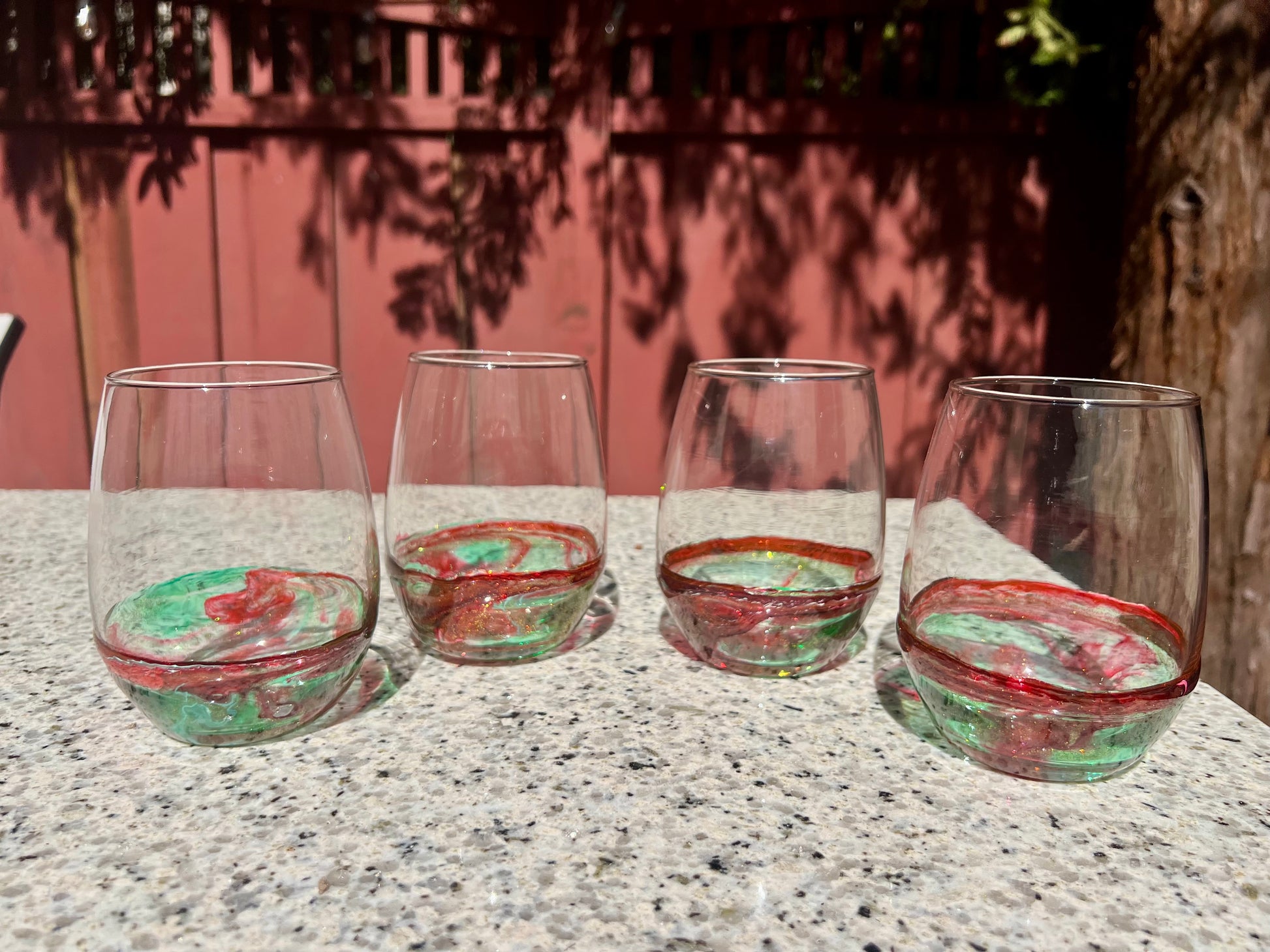 Four wine glasses with red and green swirls are arranged on a granite countertop, with a red fence and trees in the background.