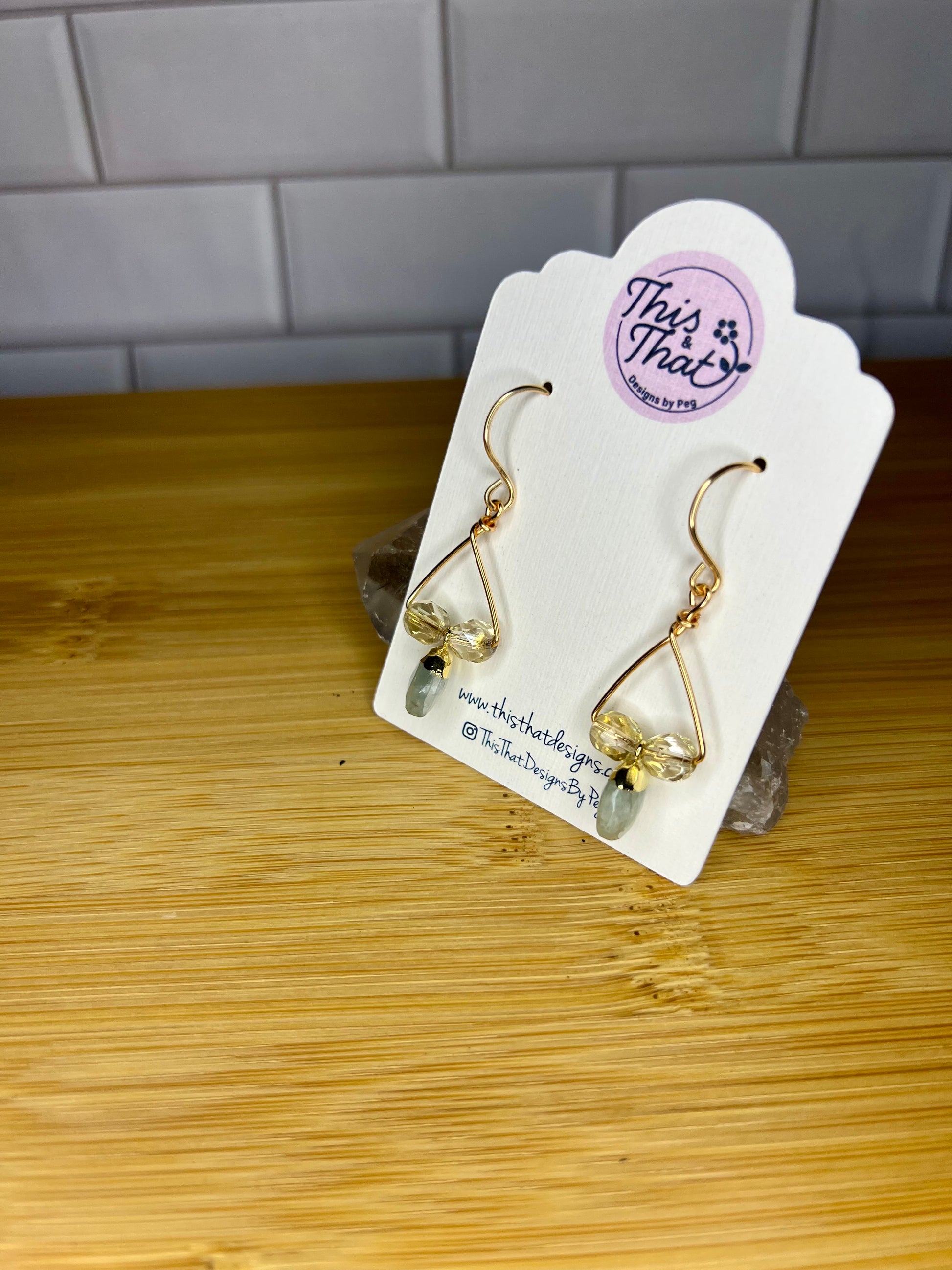 A pair of gold earrings with green stones hanging from them, displayed on a white card with a logo and text.