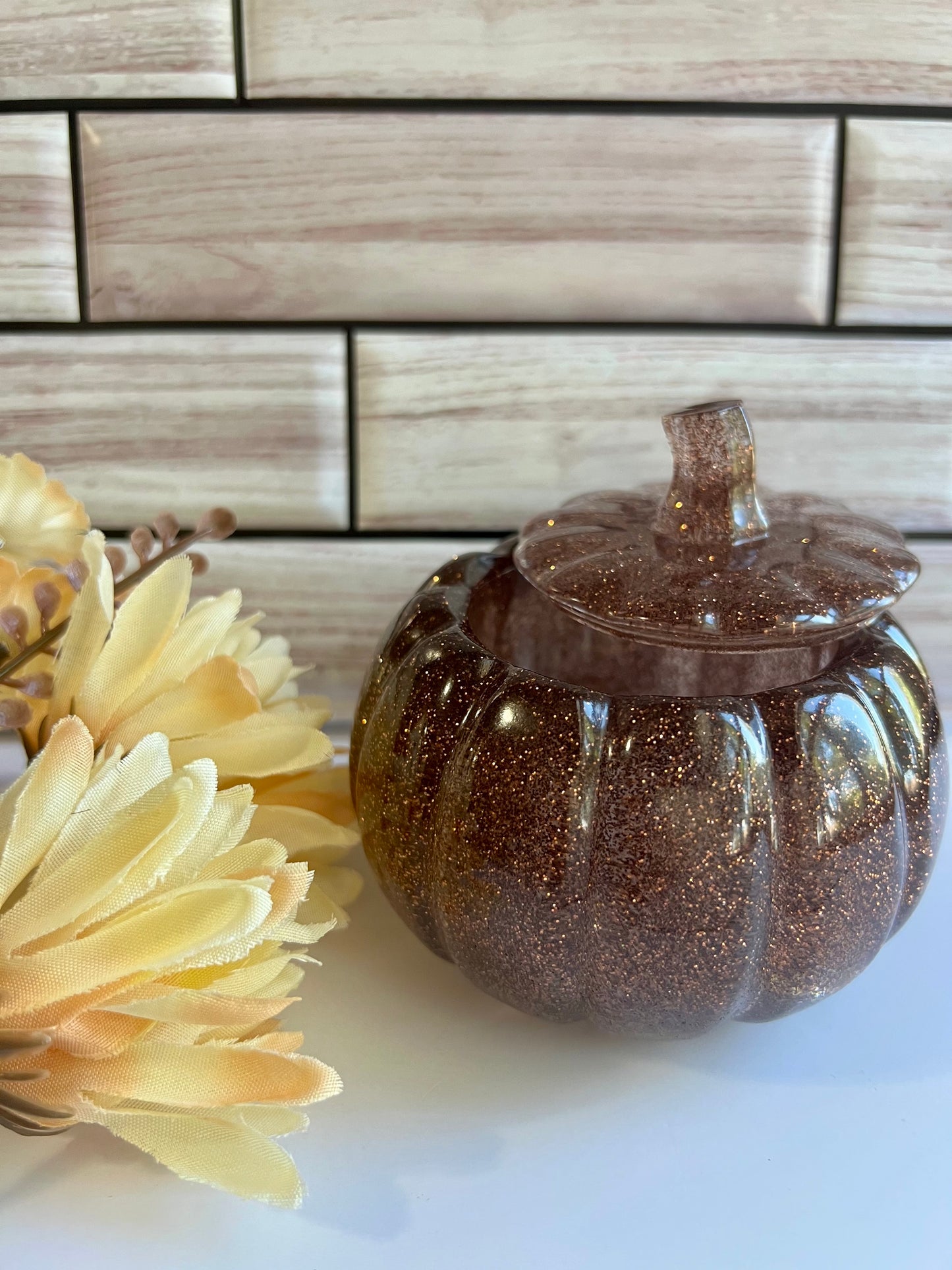 a brown glittery glass pumpkin container with a lid, placed on a white surface next to a bouquet of yellow flowers.