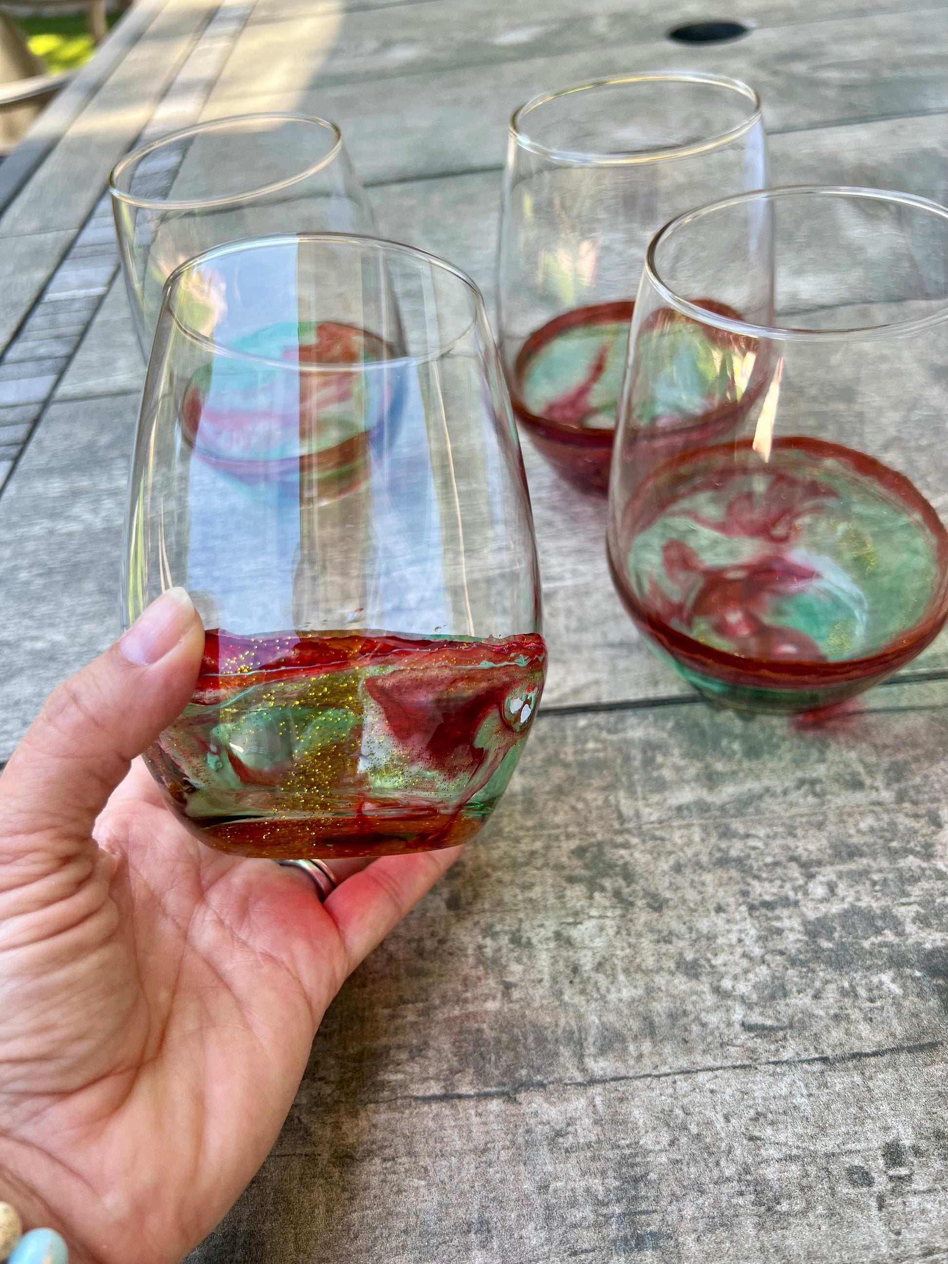 A hand holding a glass with a colorful drink, with other glasses visible in the background on a wooden table.