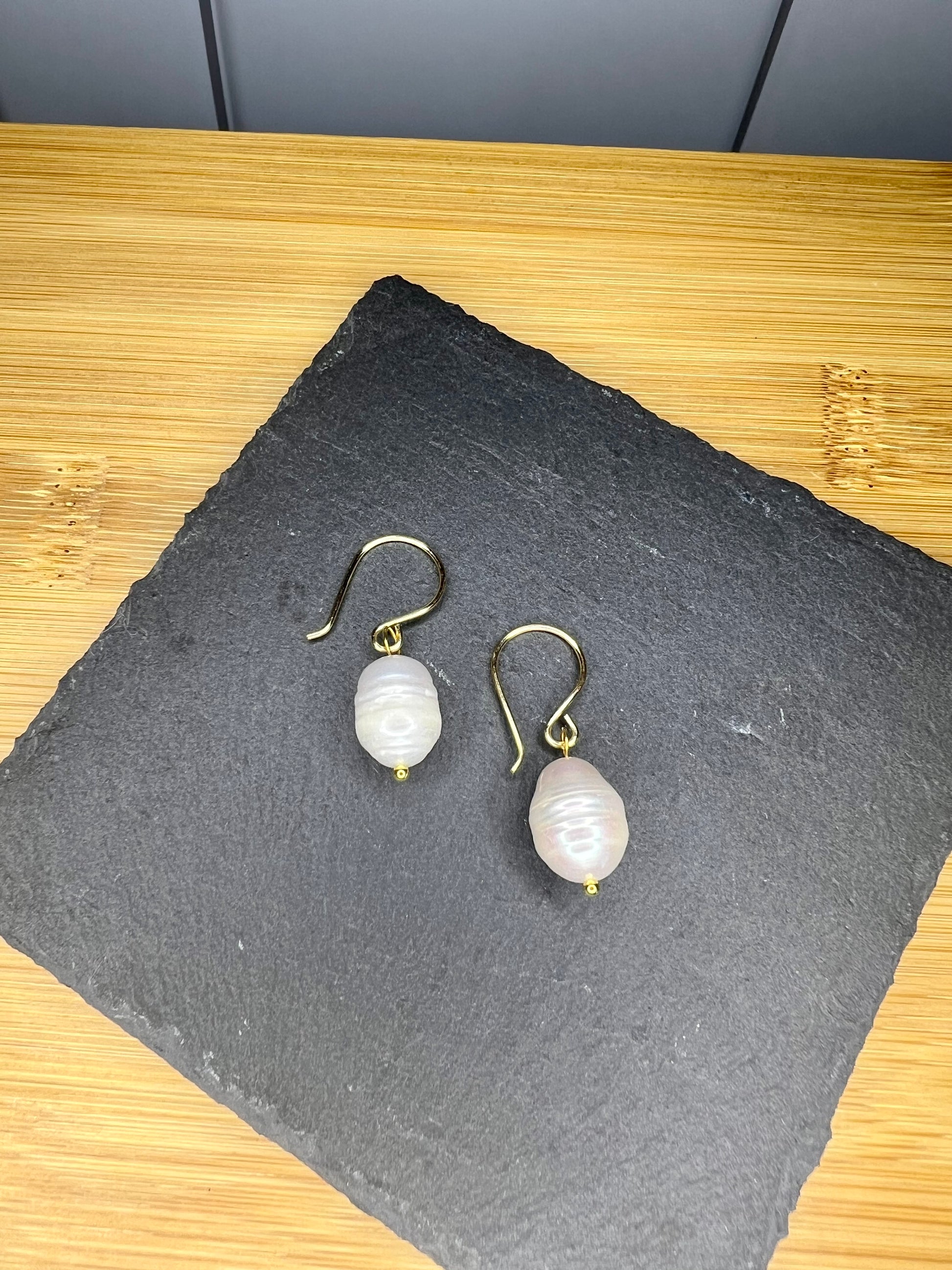 A pair of pearl earrings with a white pearl hanging from a gold hook rests on a black slate.