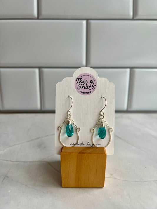 A pair of earrings with turquoise stones, displayed on a wooden stand against a white tiled wall.