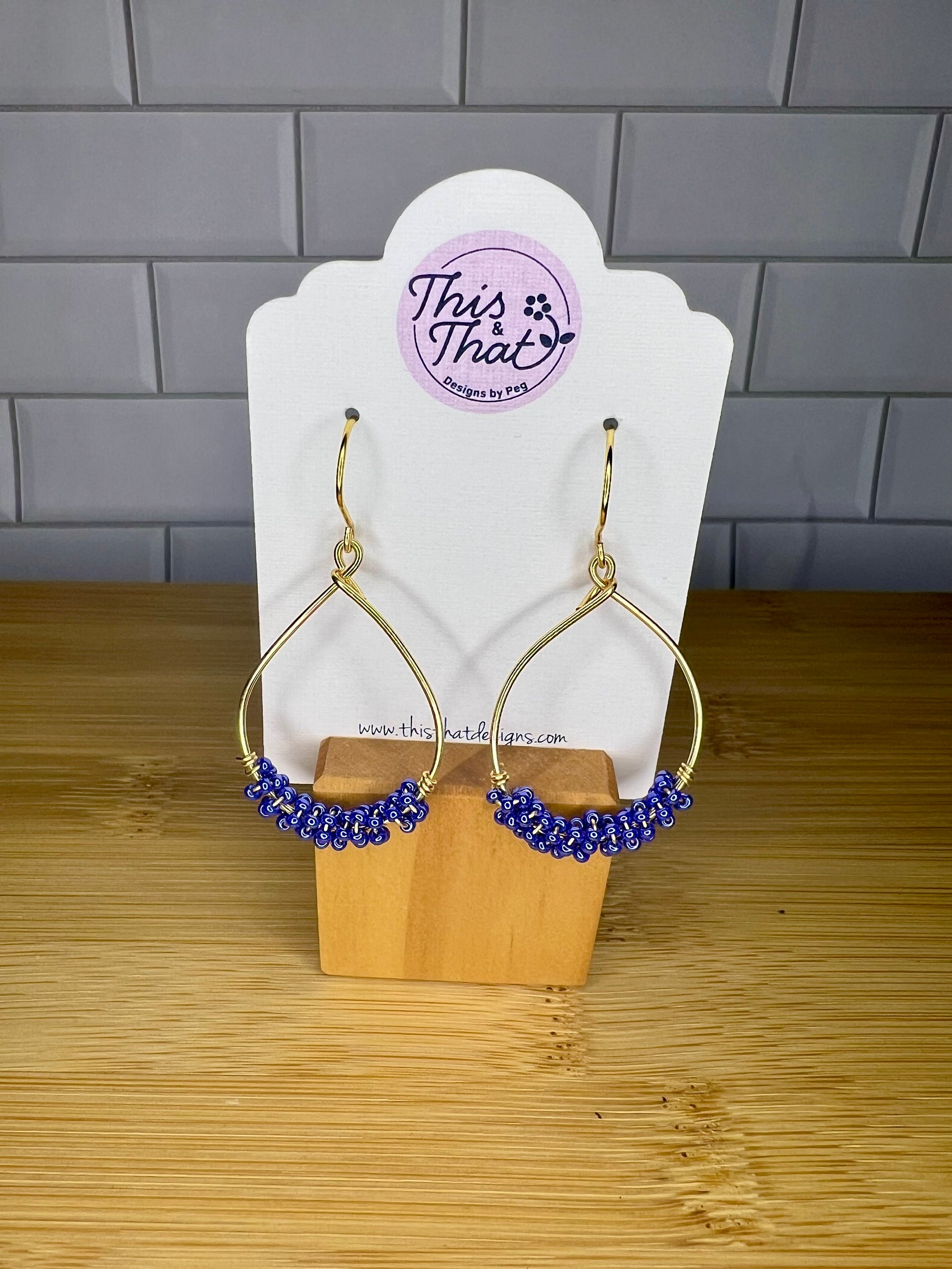 A pair of gold earrings with blue beads, displayed on a wooden stand with a white card featuring the text "This That Designs by PGB".