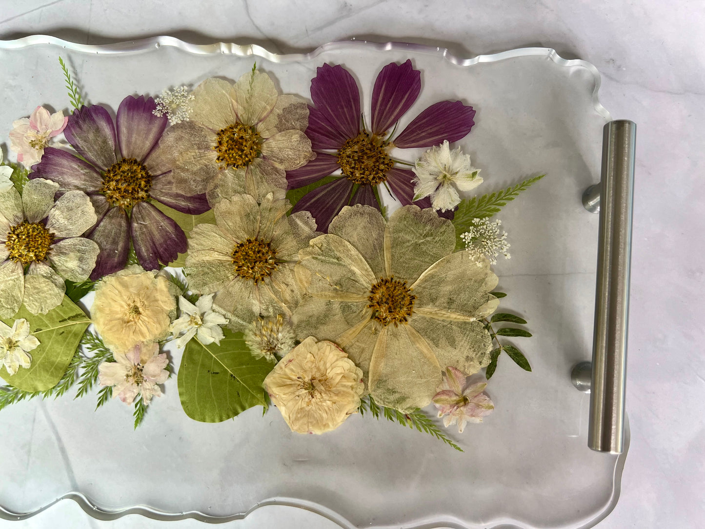A clear glass tray with a metal handle, displaying a variety of colorful dried flowers and leaves arranged on it.