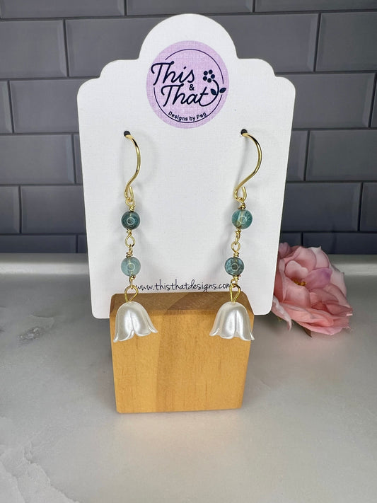 A pair of earrings with white flower-shaped charms and green gemstone accents, displayed on a wooden stand.