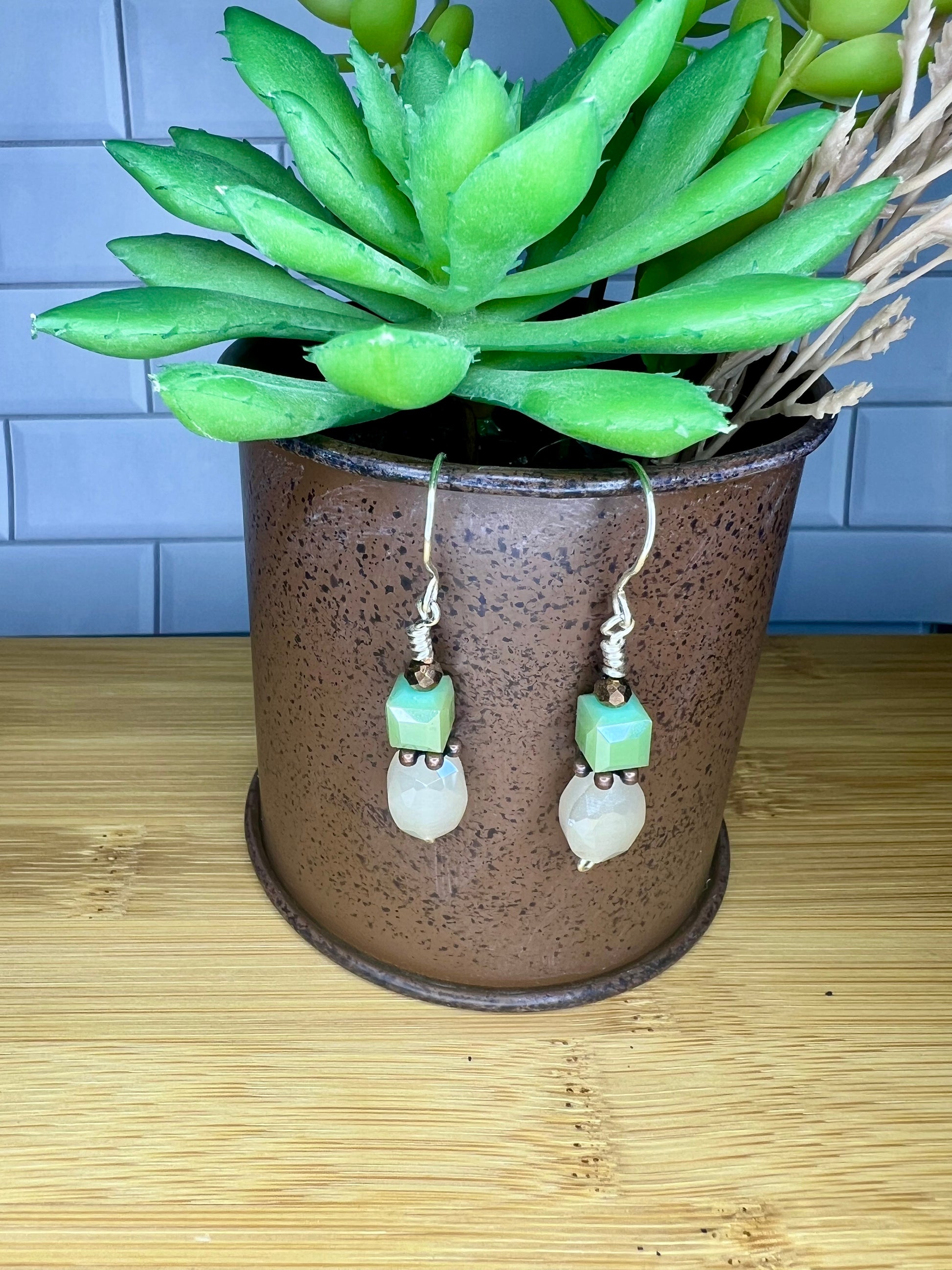 A pair of earrings with green and white beads hanging from a brown pot, placed on a wooden surface.