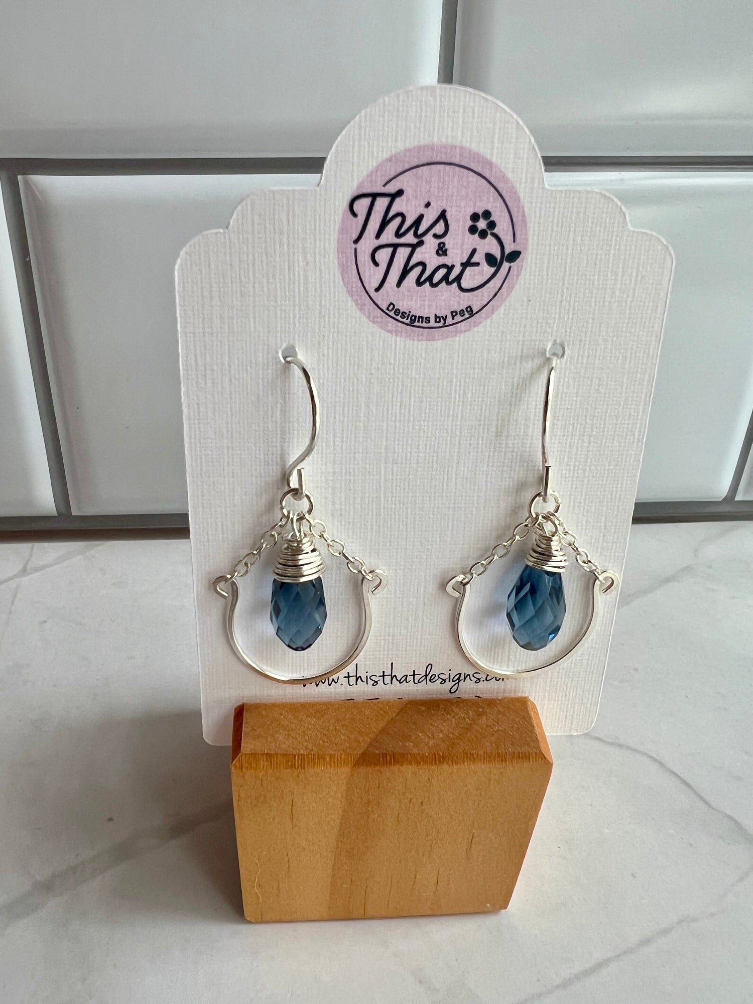 This image shows a pair of earrings on a white display stand. The earrings have a blue crystal design and are hanging from a metal hook. The display stand has a wooden base and a circular logo on it.