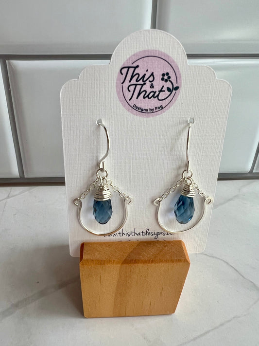 This image shows a pair of earrings on a white display stand. The earrings have a blue crystal design and are hanging from a metal hook. The display stand has a wooden base and a circular logo on it.