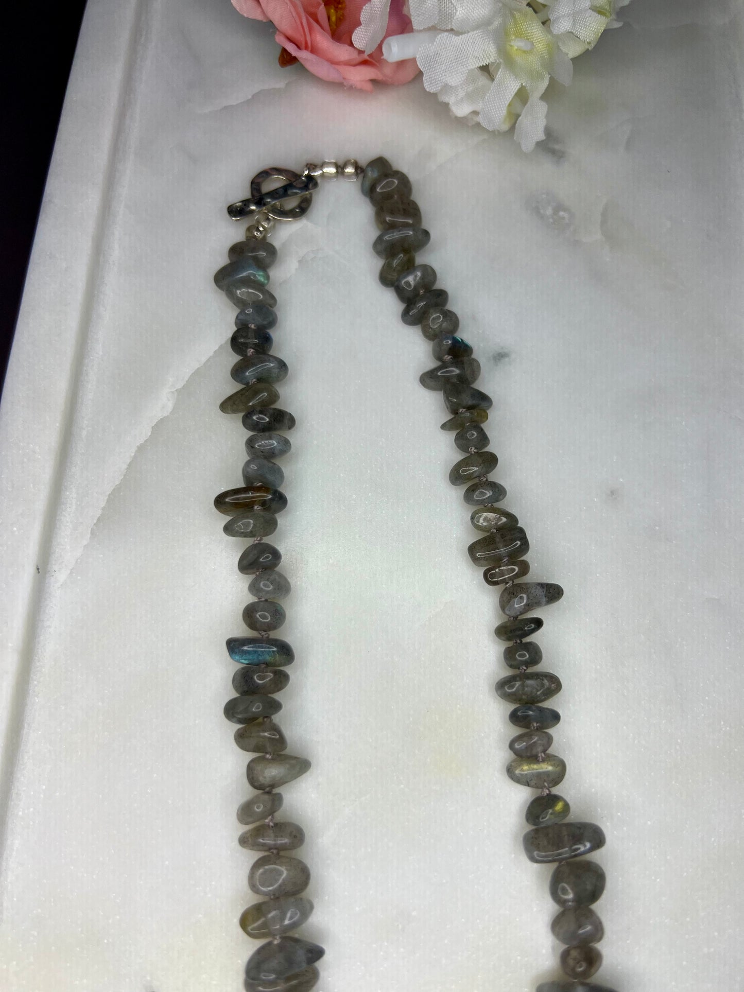 A necklace made of small, irregularly shaped gray stones is displayed on a white surface, with a pink flower in the background.