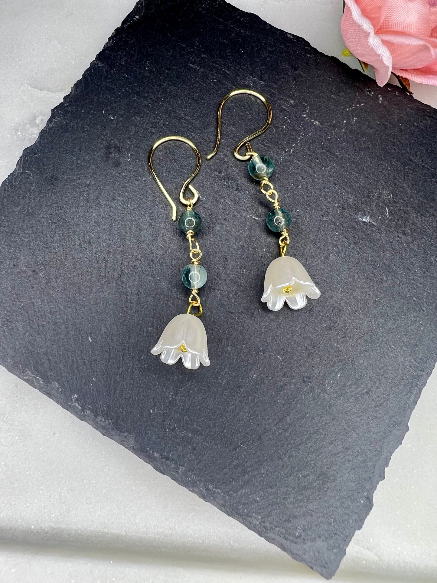 A pair of elegant earrings with white petals and green gemstones, resting on a dark surface.