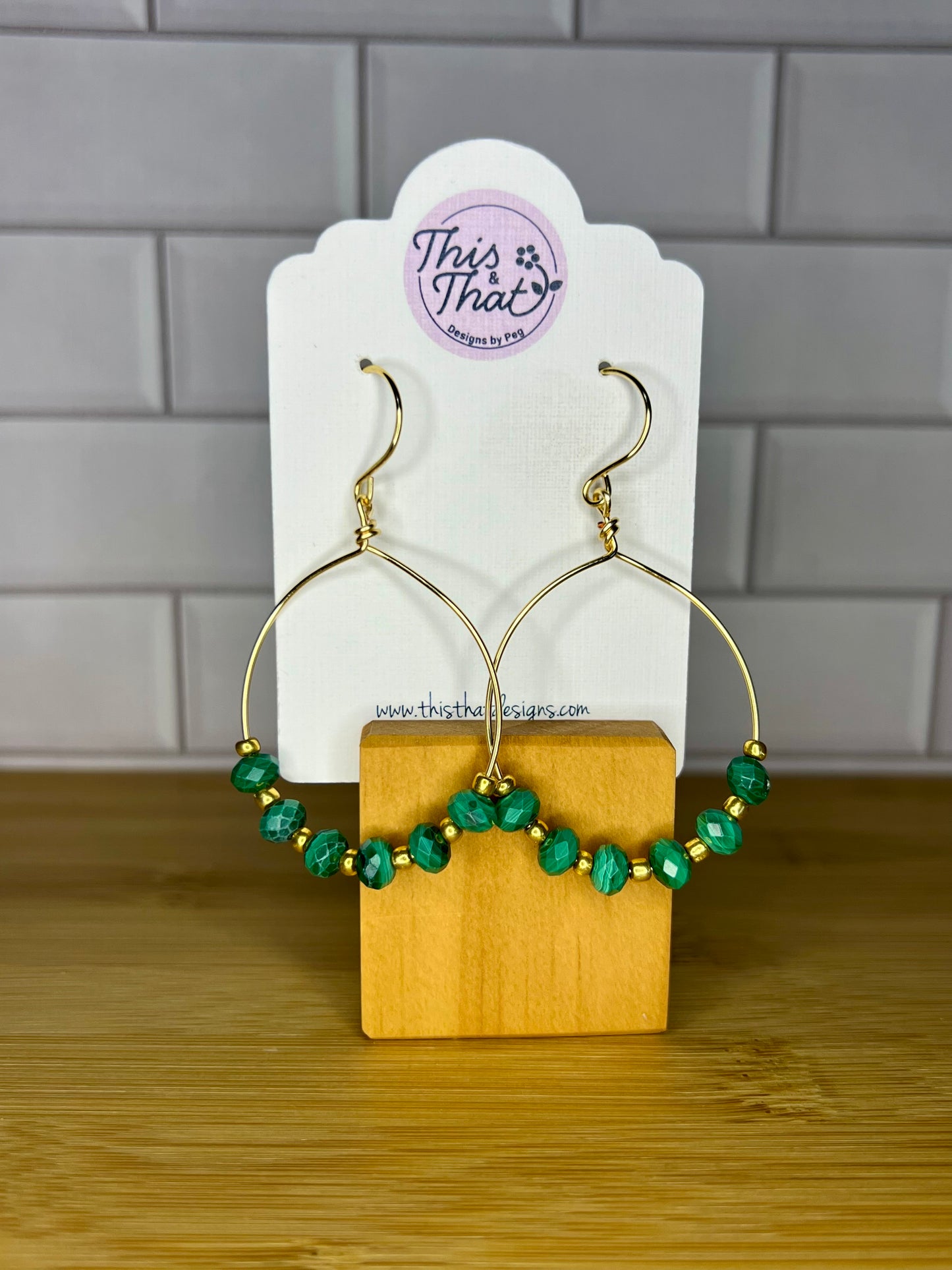 A pair of gold hoop earrings with green gemstone beads, displayed on a wooden stand against a tiled wall.