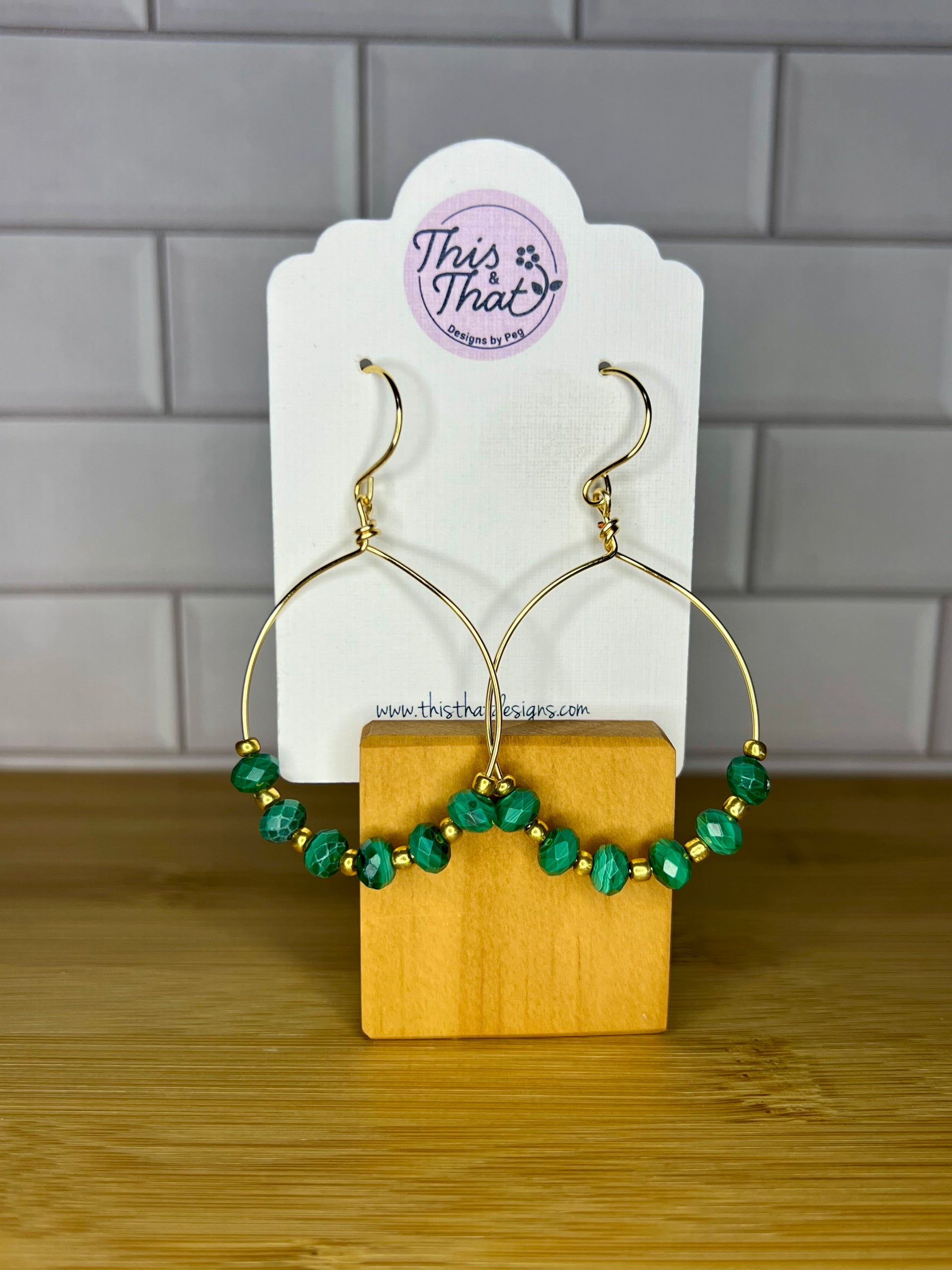 A pair of gold hoop earrings with green gemstone beads, displayed on a wooden stand against a tiled wall.