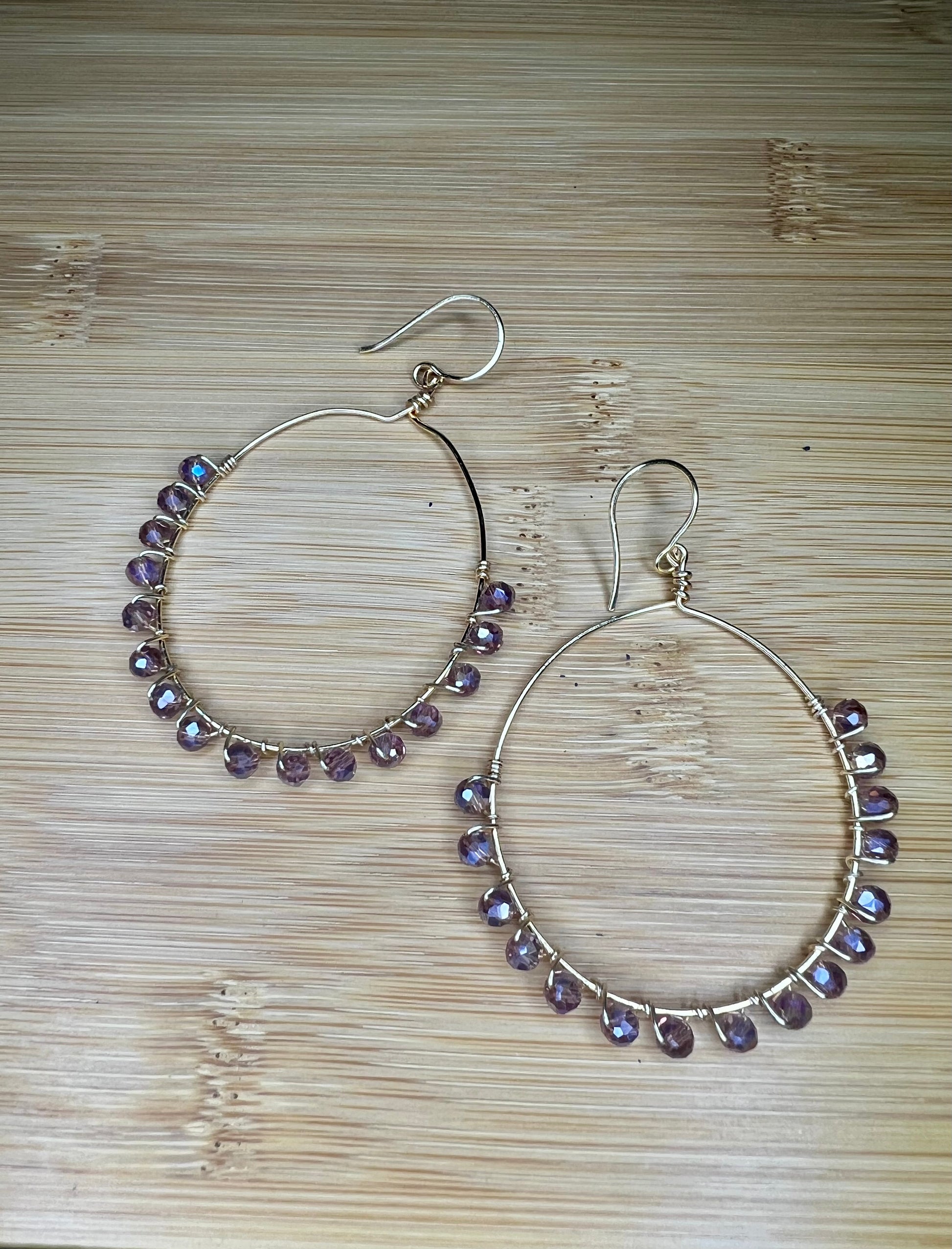 A pair of silver hoop earrings with purple gemstone beads hanging from them, placed on a wooden surface.