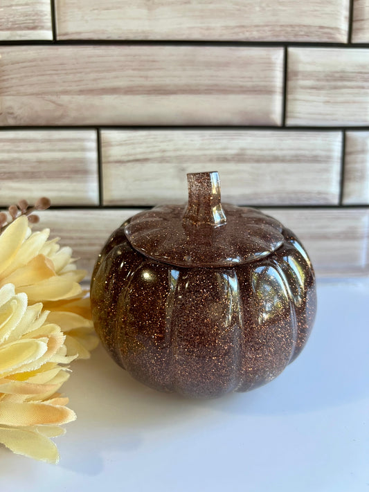 A brown glittery glass pumpkin-shaped container sits on a white surface, with a yellow flower to the left.