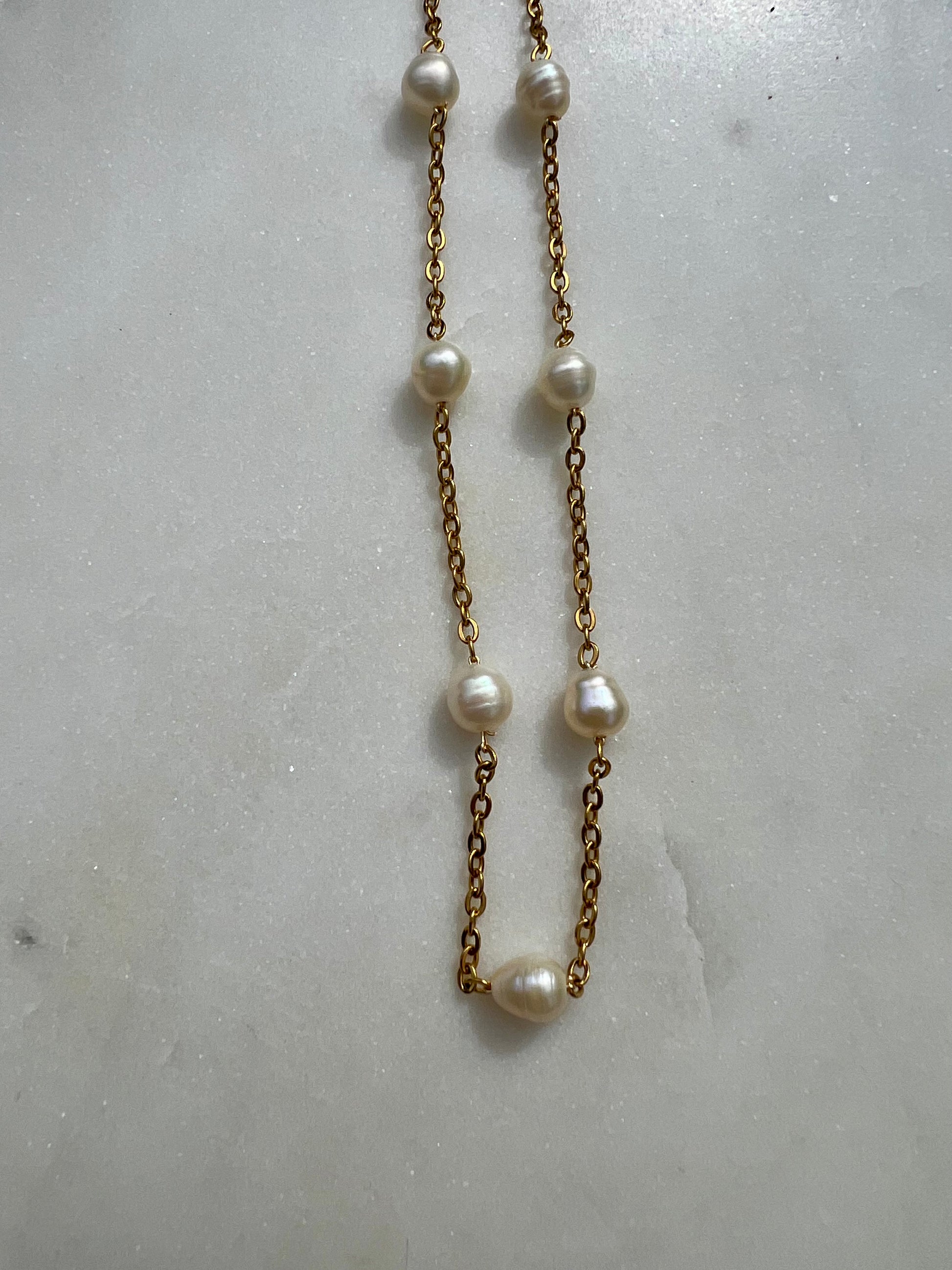 A necklace with a series of white pearls and a gold chain is displayed against a white marble background.