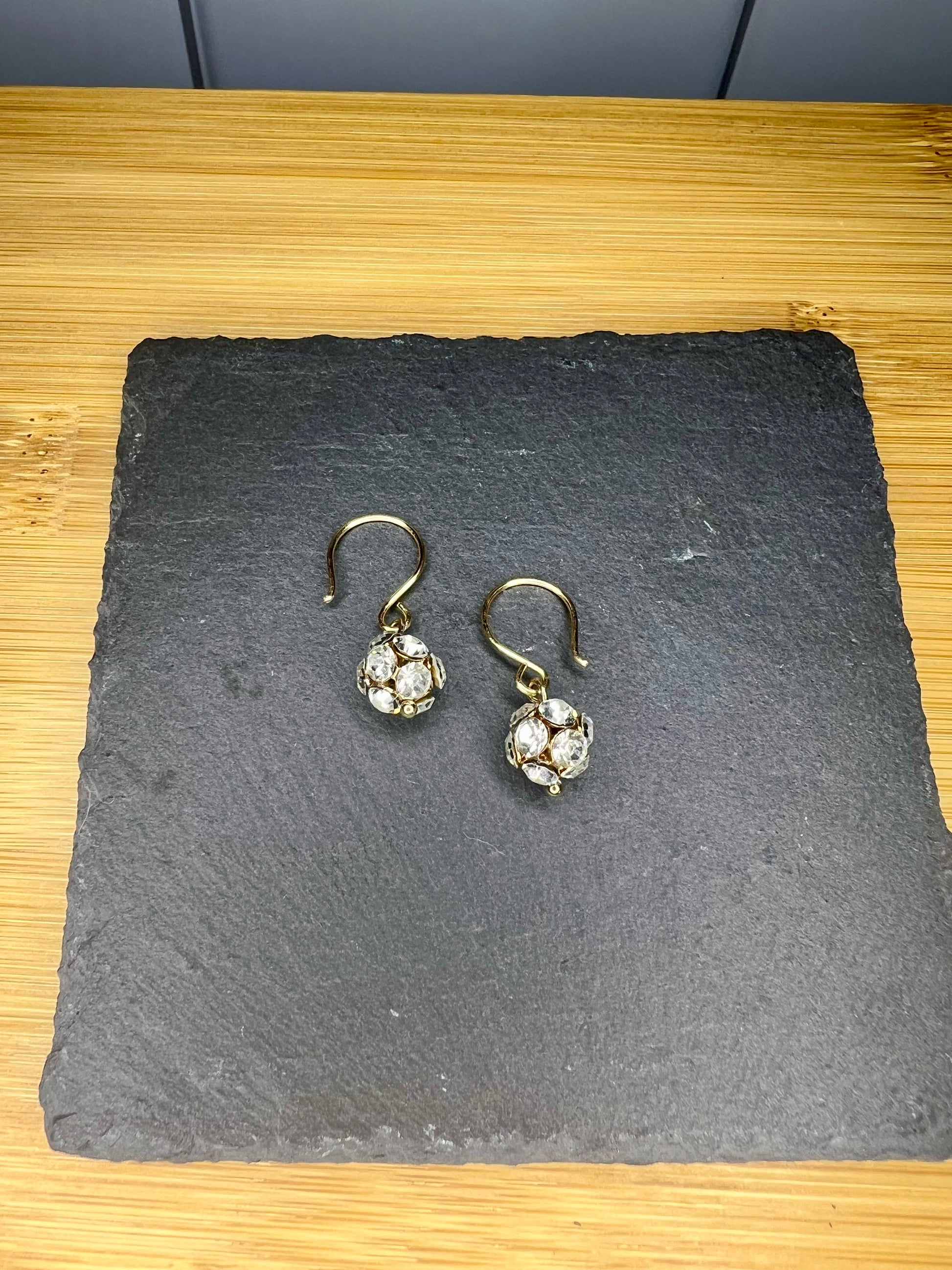 A pair of gold earrings with small, clear stones hanging from them, placed on a dark slate or slate-like surface.