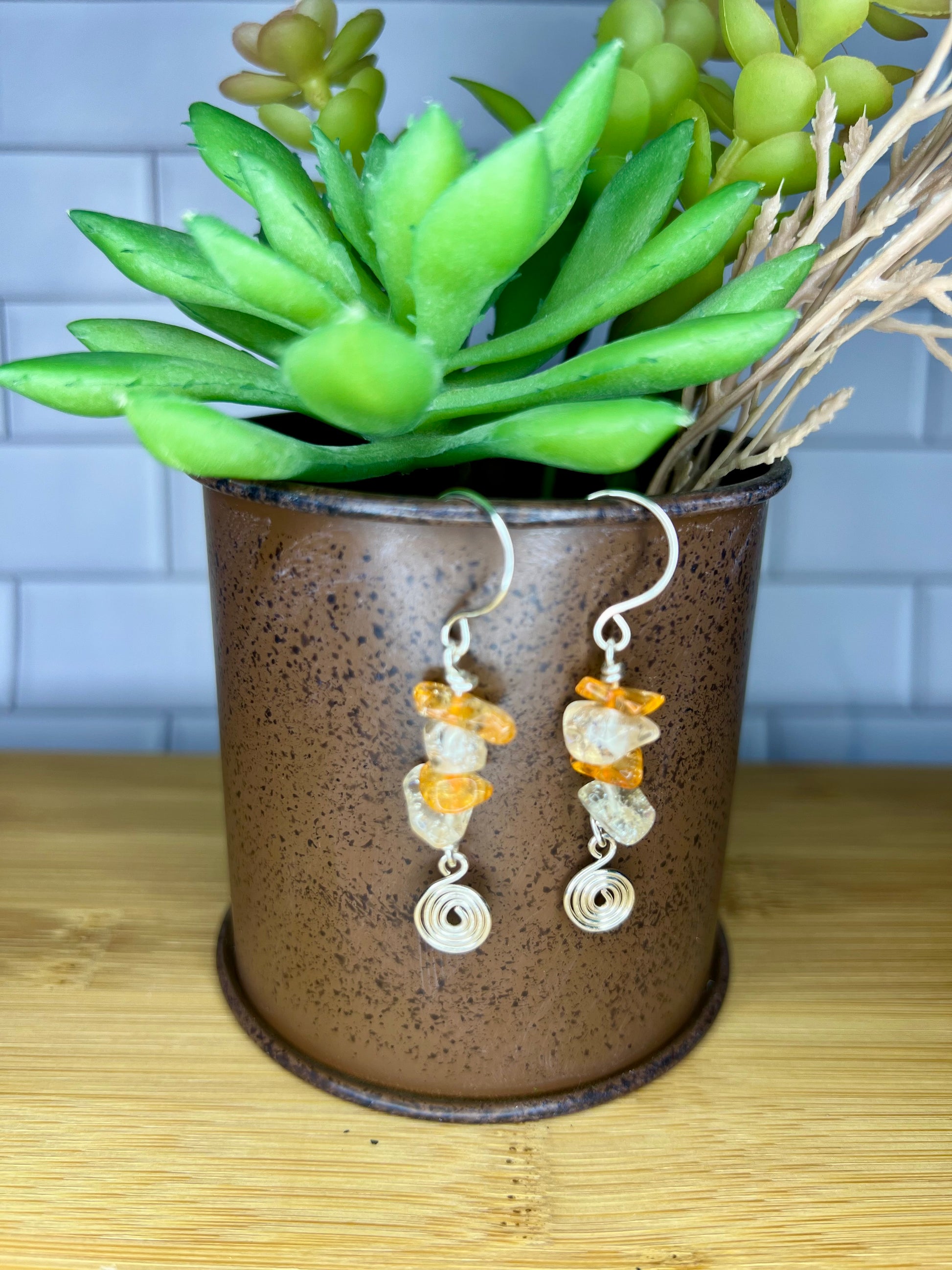 A pair of earrings with orange and white stones hanging from a brown metal container, placed on a wooden surface.