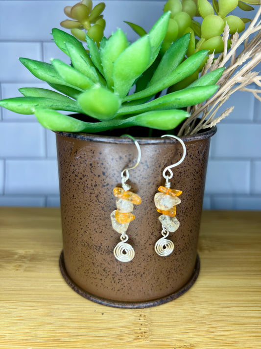 A pair of earrings with orange and white stones hanging from a brown metal container, placed on a wooden surface.