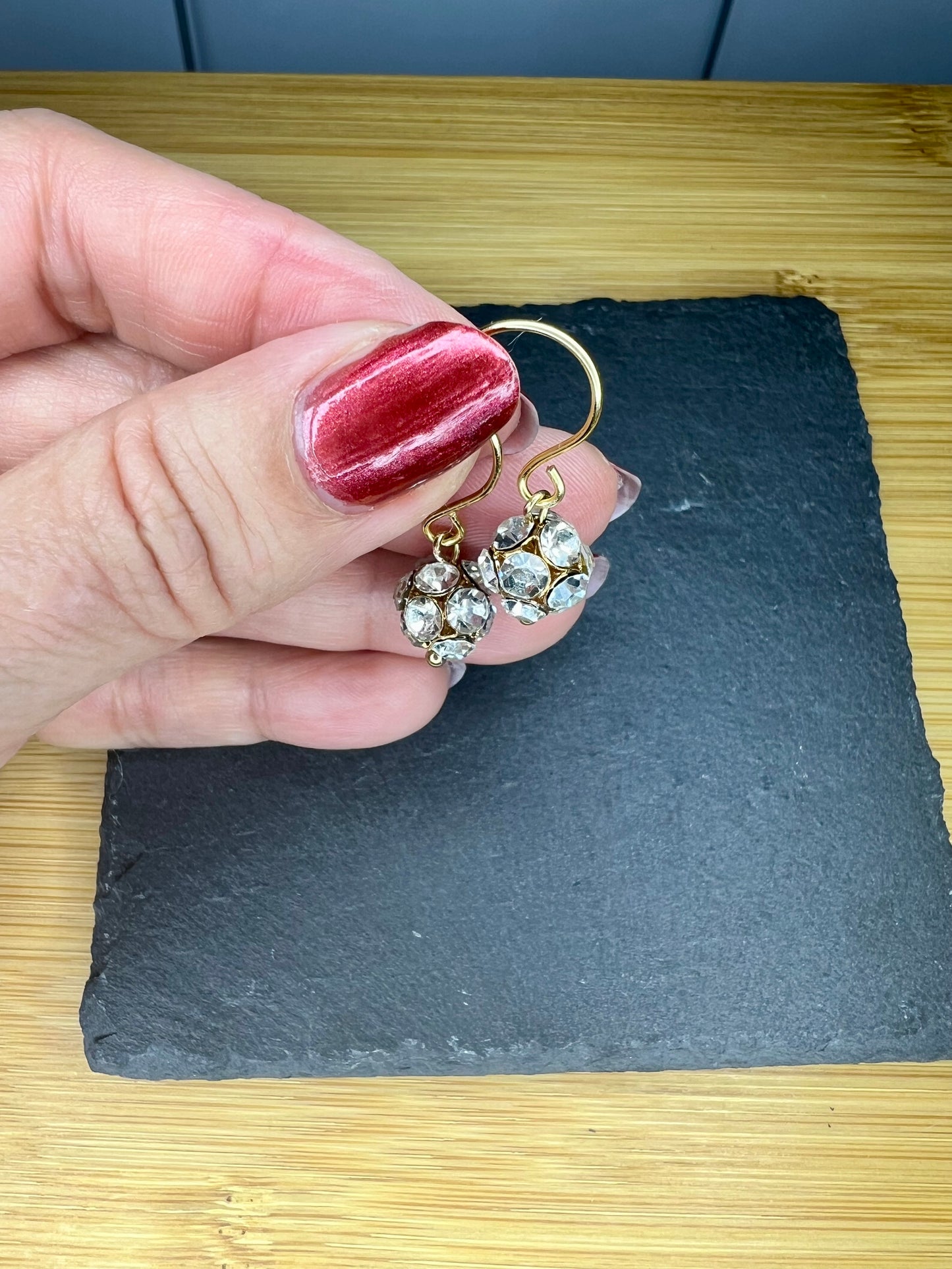 A hand holding a pair of diamond earrings with a red nail polish on the thumb.
