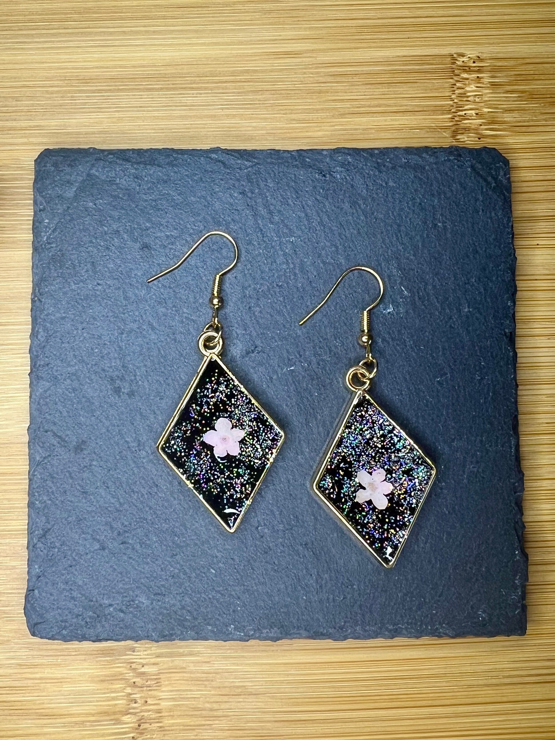 A pair of diamond-shaped earrings with a glittery, star-like design and a small pink flower in the center, placed on a dark slate surface.