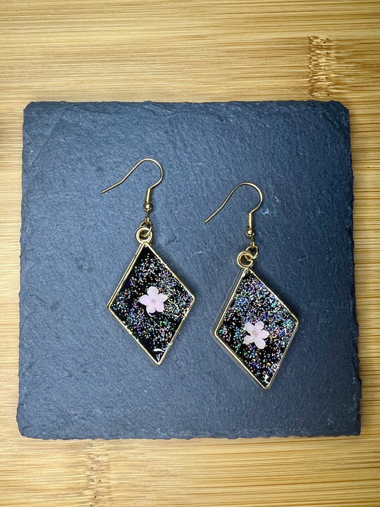 A pair of diamond-shaped earrings with a glittery, star-like design and a small pink flower in the center, placed on a dark slate surface.