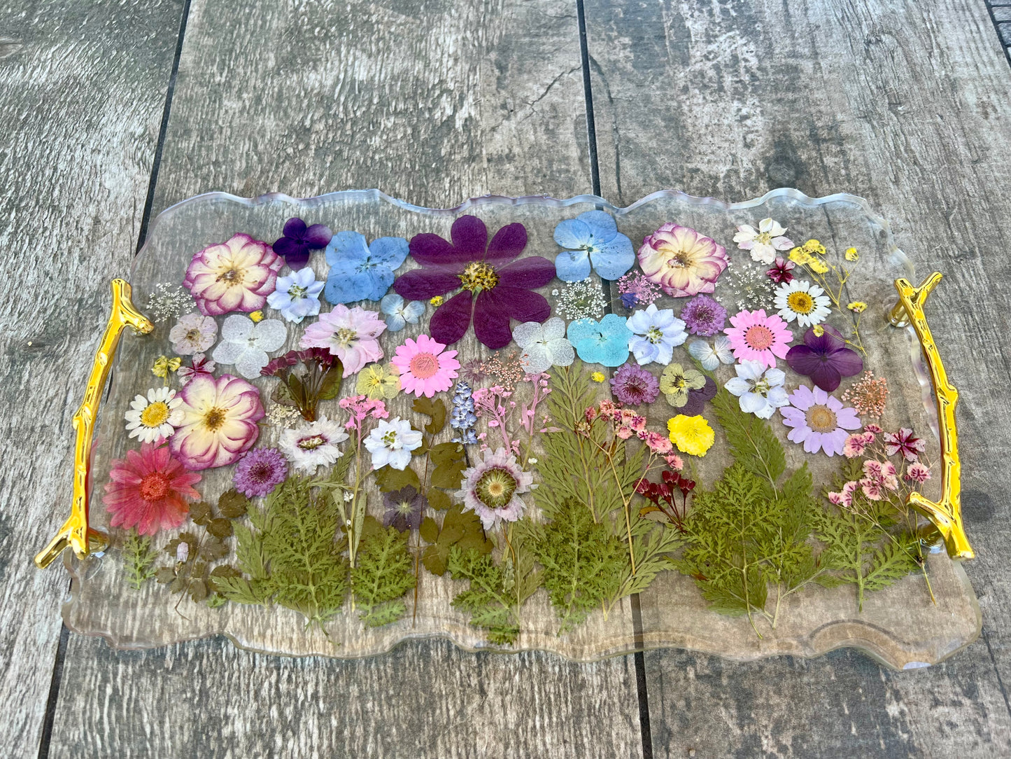 A clear glass tray with a gold handle, filled with a variety of colorful flowers and leaves, placed on a wooden surface.