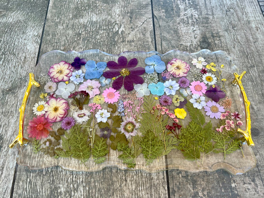 A clear glass tray with a gold handle, filled with a variety of colorful flowers and leaves, placed on a wooden surface.