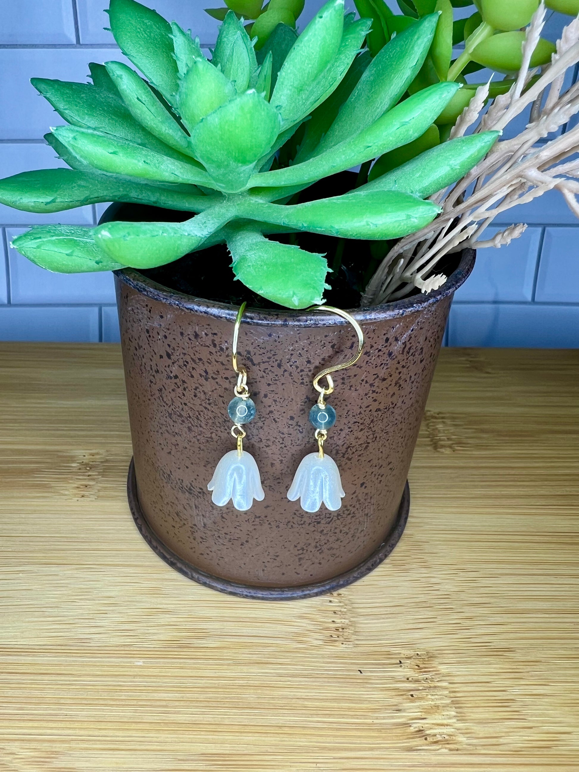 A pair of white earrings with green leaves hanging from them, placed in a brown pot with a potted plant.