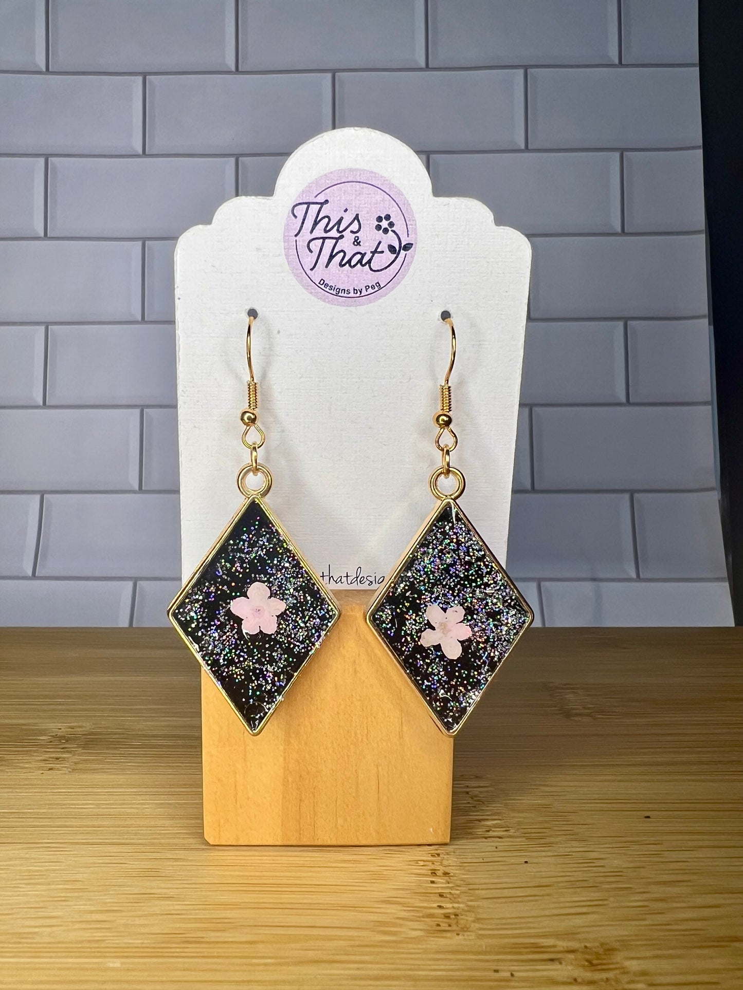 A pair of diamond-shaped earrings with a glittery black background and a pink flower design, displayed on a wooden stand with a white tag.