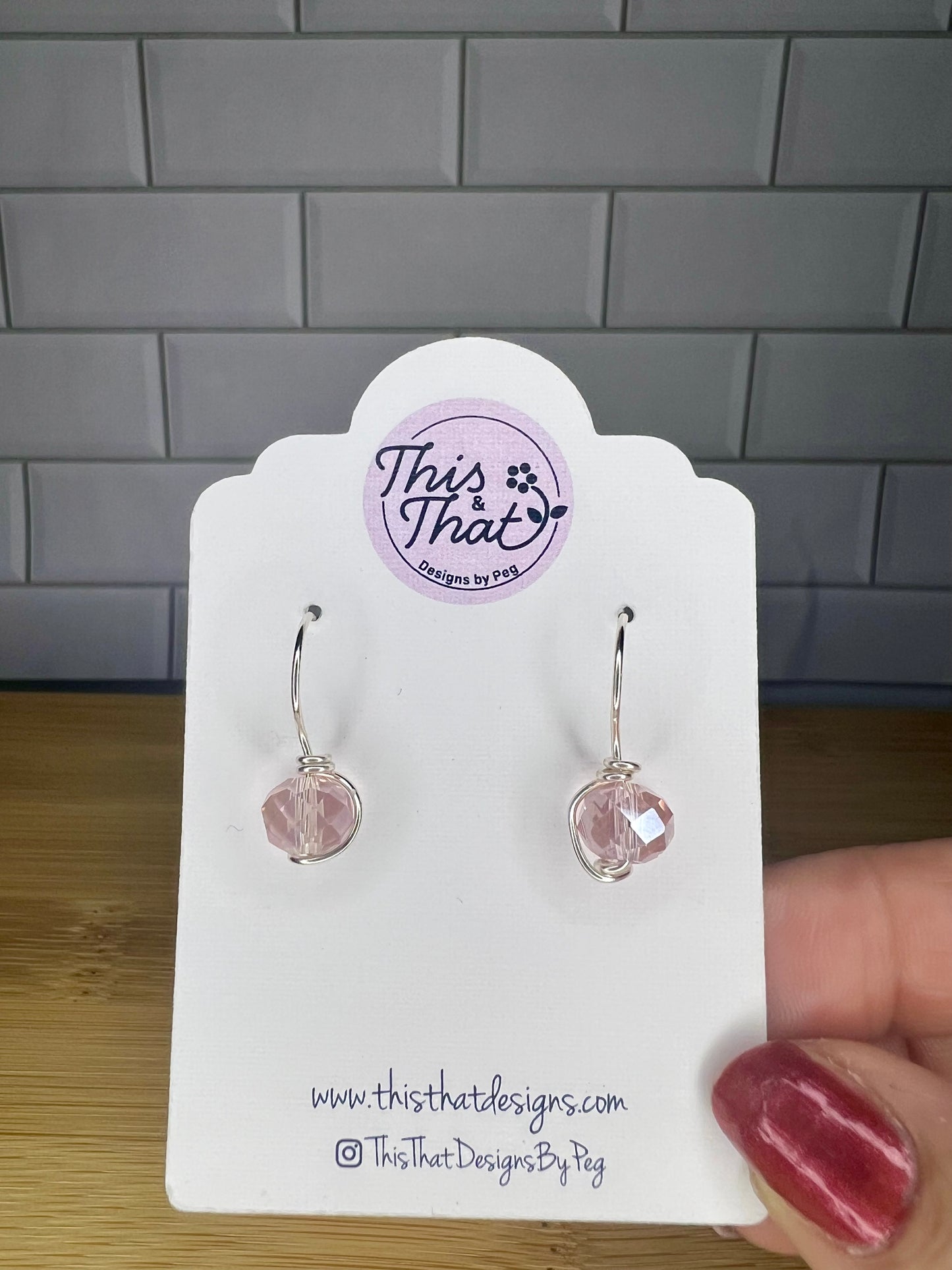 a person holding a white earring card with a pair of earrings attached to it. The earring card has a website address and a social media handle printed on it.