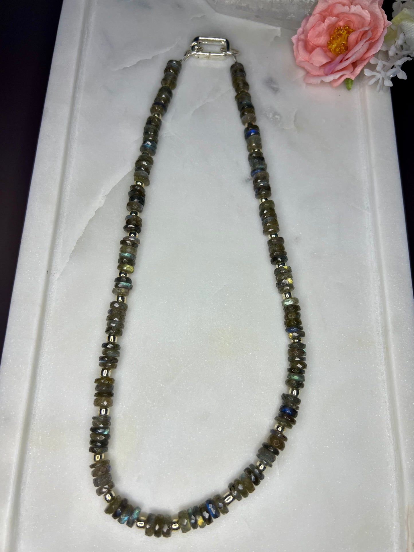 A necklace made of small, multicolored beads is displayed on a white surface, with a pink flower resting on the right side of the image.