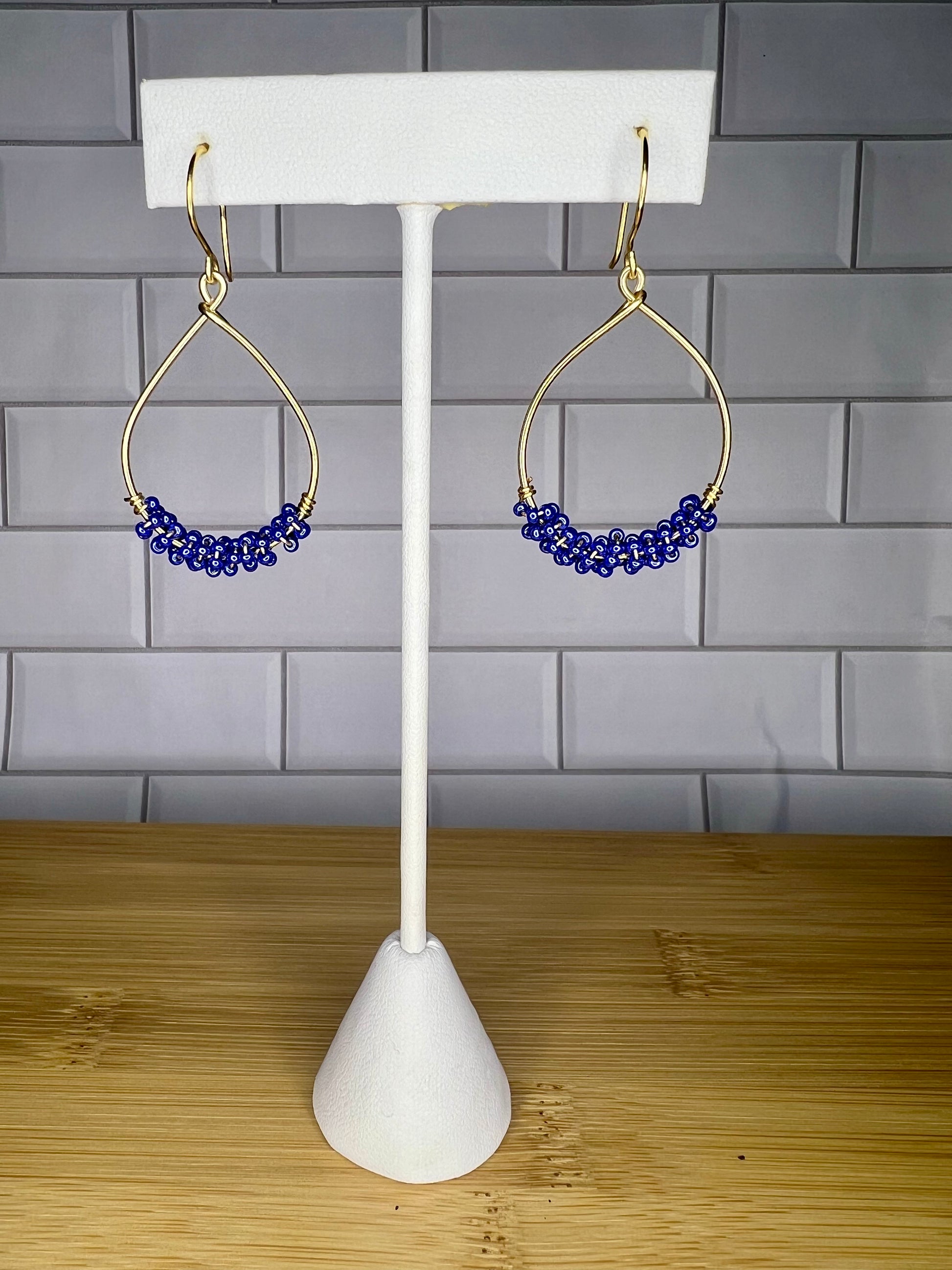 A pair of gold earrings with blue beads hanging from them, displayed on a white stand against a gray tiled wall.