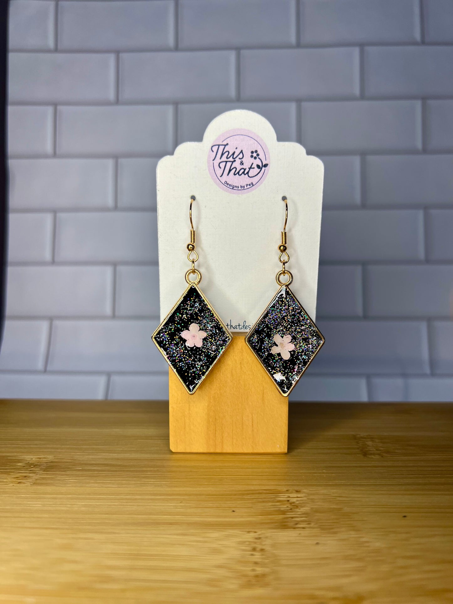 A pair of diamond-shaped earrings with a glittery black and white design, displayed on a wooden stand against a tiled wall background.