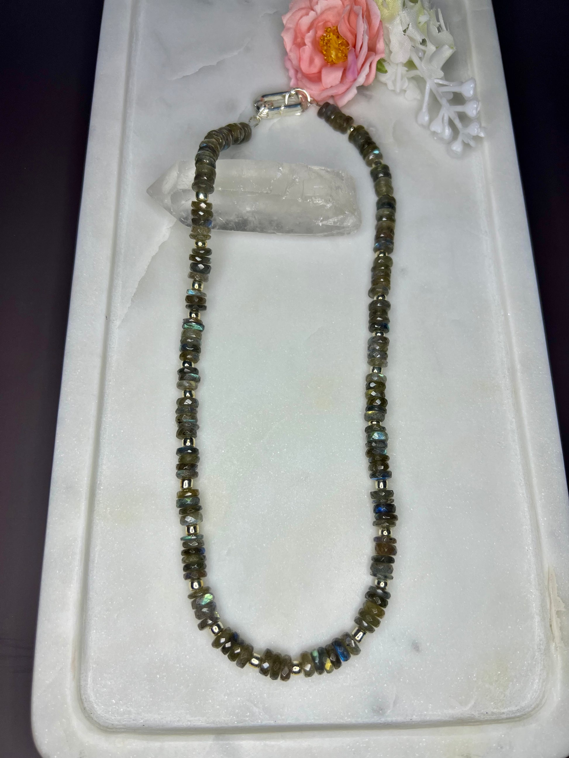 a necklace with a long chain of small, square-shaped beads in various shades of brown and gray, resting on a white marble surface. A pink flower is also present in the background.