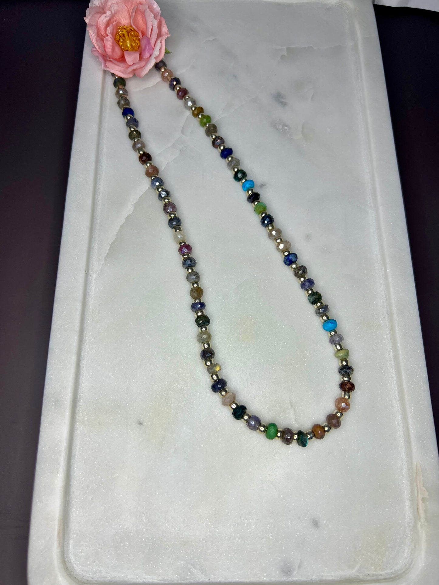 A necklace with multicolored beads is displayed on a white marble surface, accompanied by a single pink flower.