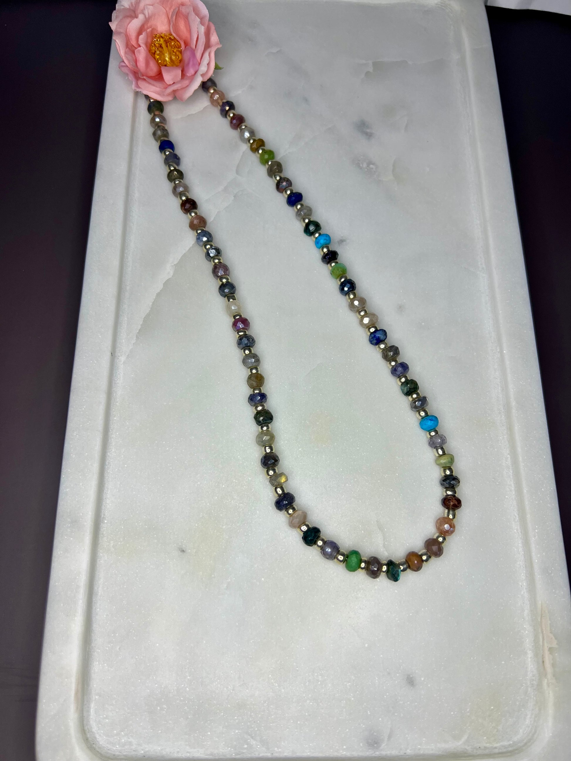 A necklace with multicolored beads is displayed on a white marble surface, accompanied by a single pink flower.