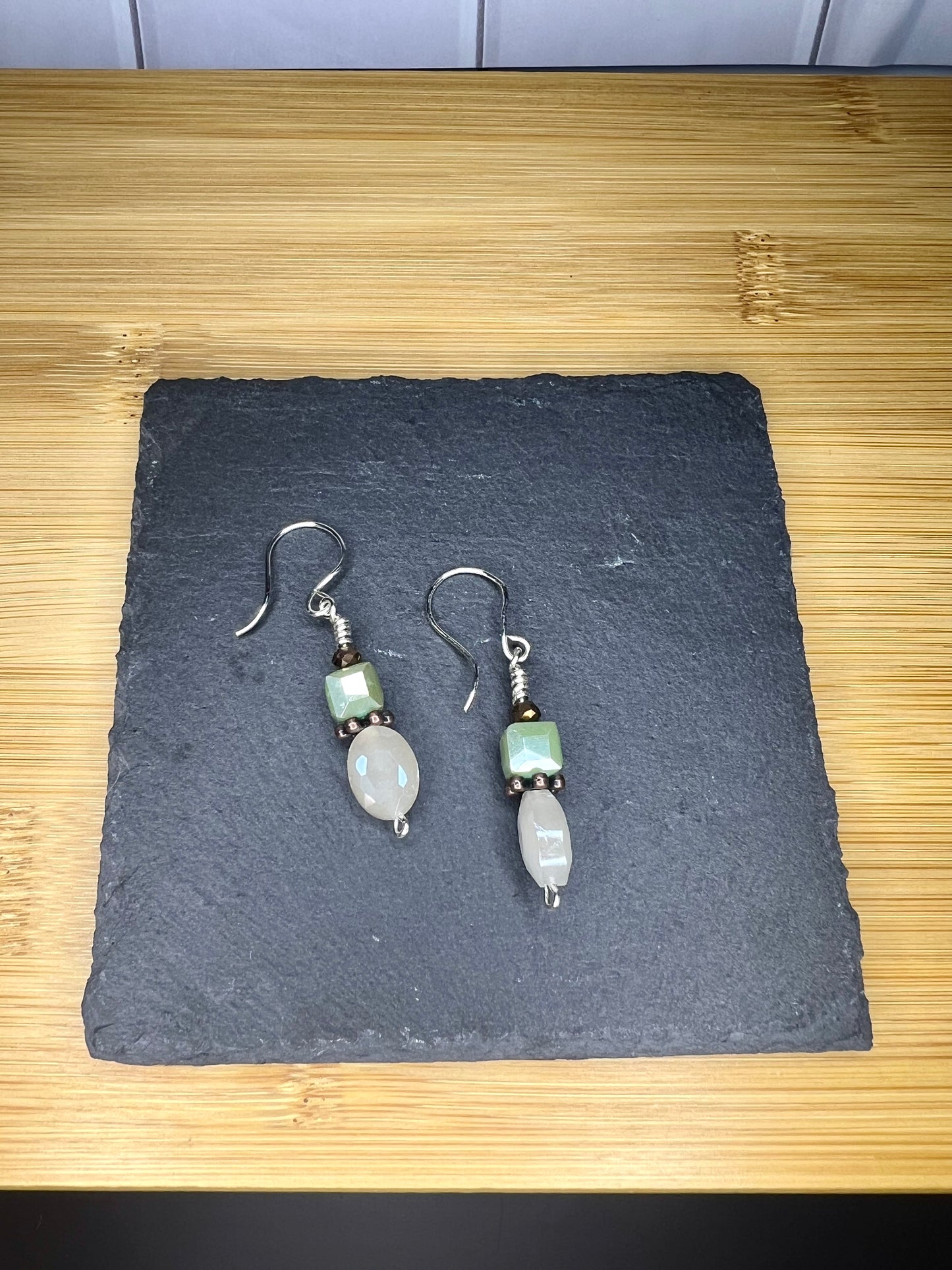 A pair of silver earrings with green stones and a small smiley face charm, placed on a black slate or slate board.
