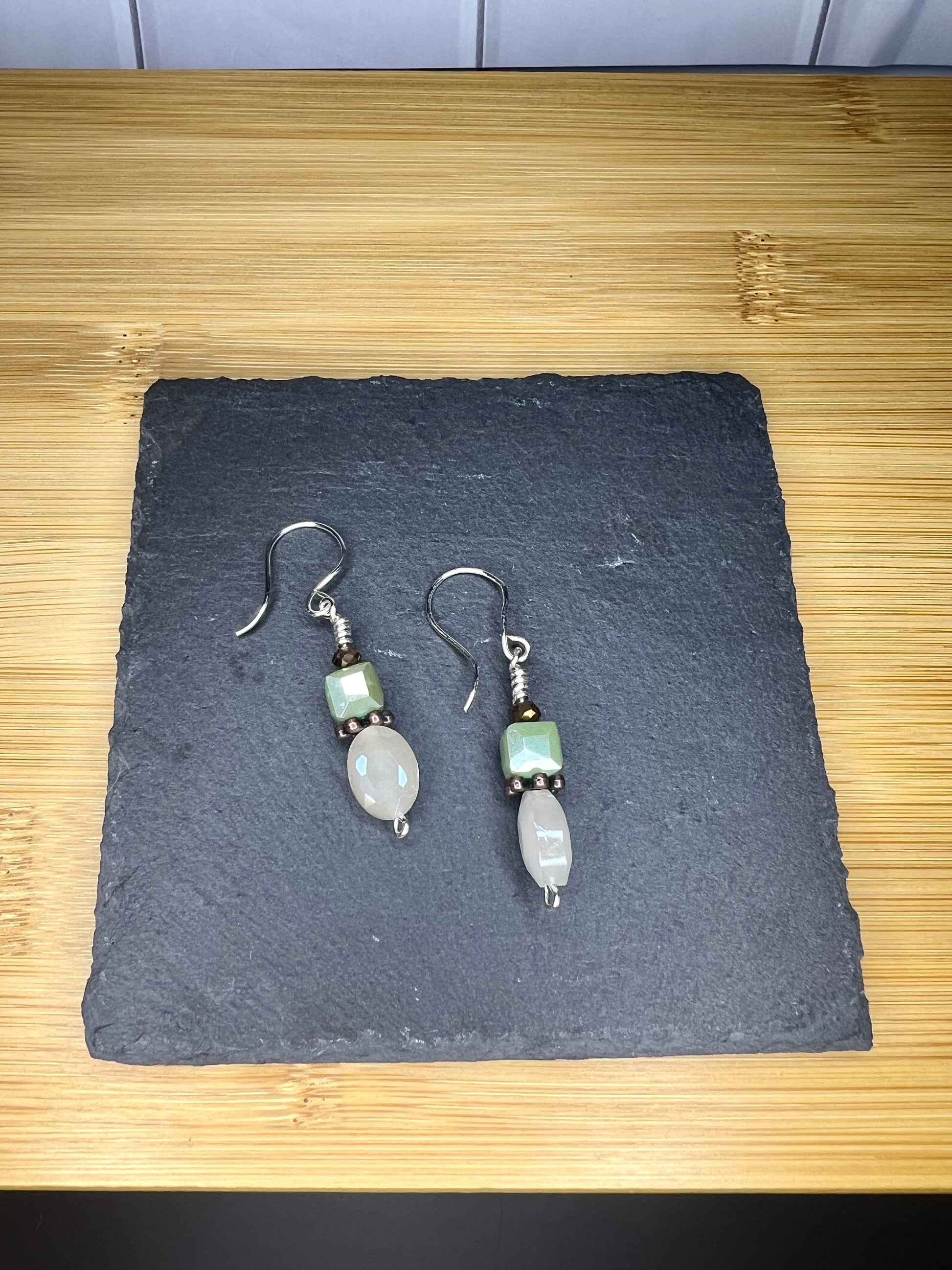 A pair of silver earrings with green stones and a small smiley face charm, placed on a black slate or slate board.